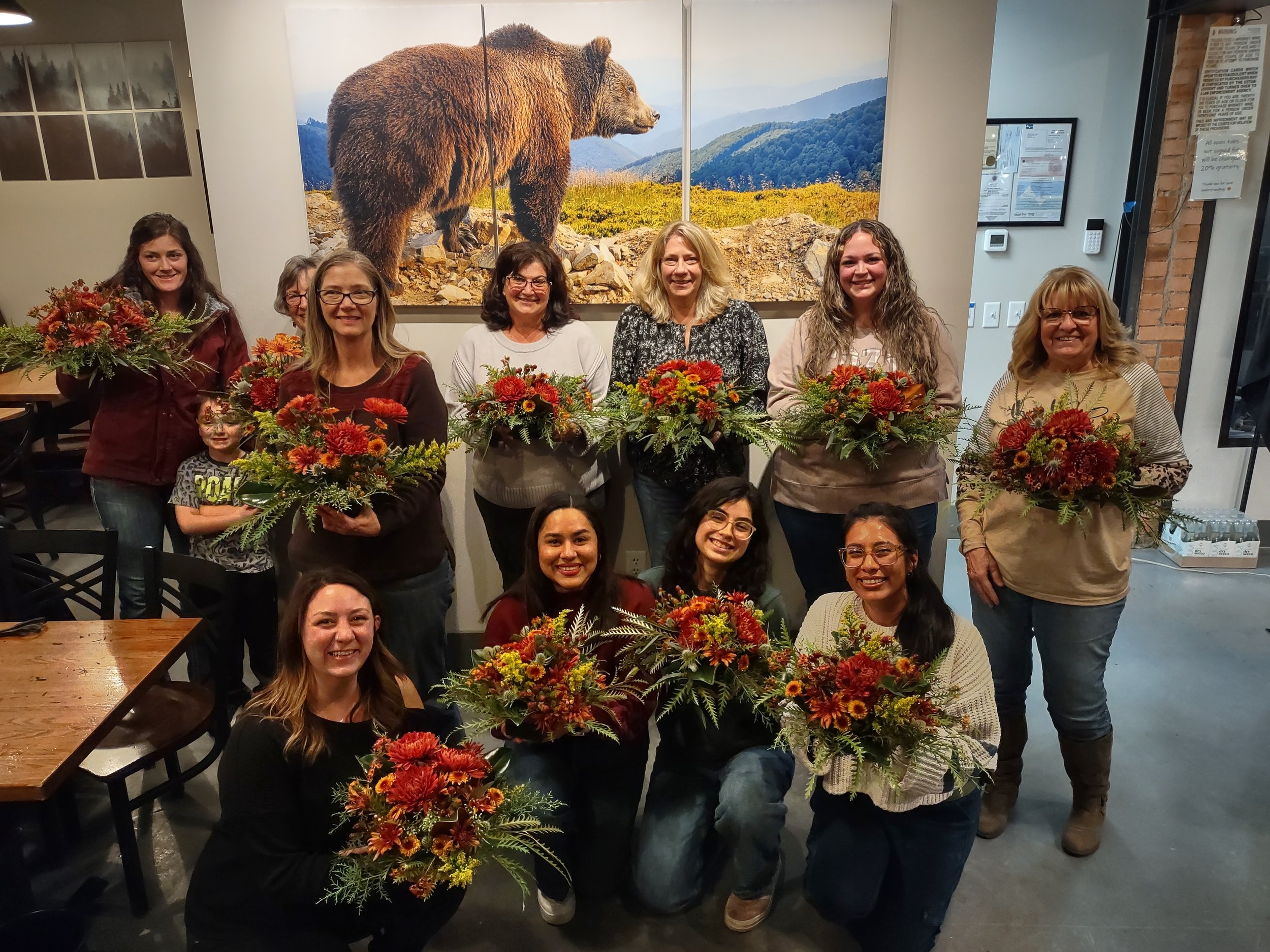 Group of women and children posing indoors with fall flower arrangements, standing in front of a large mountain landscape poster with a bear.