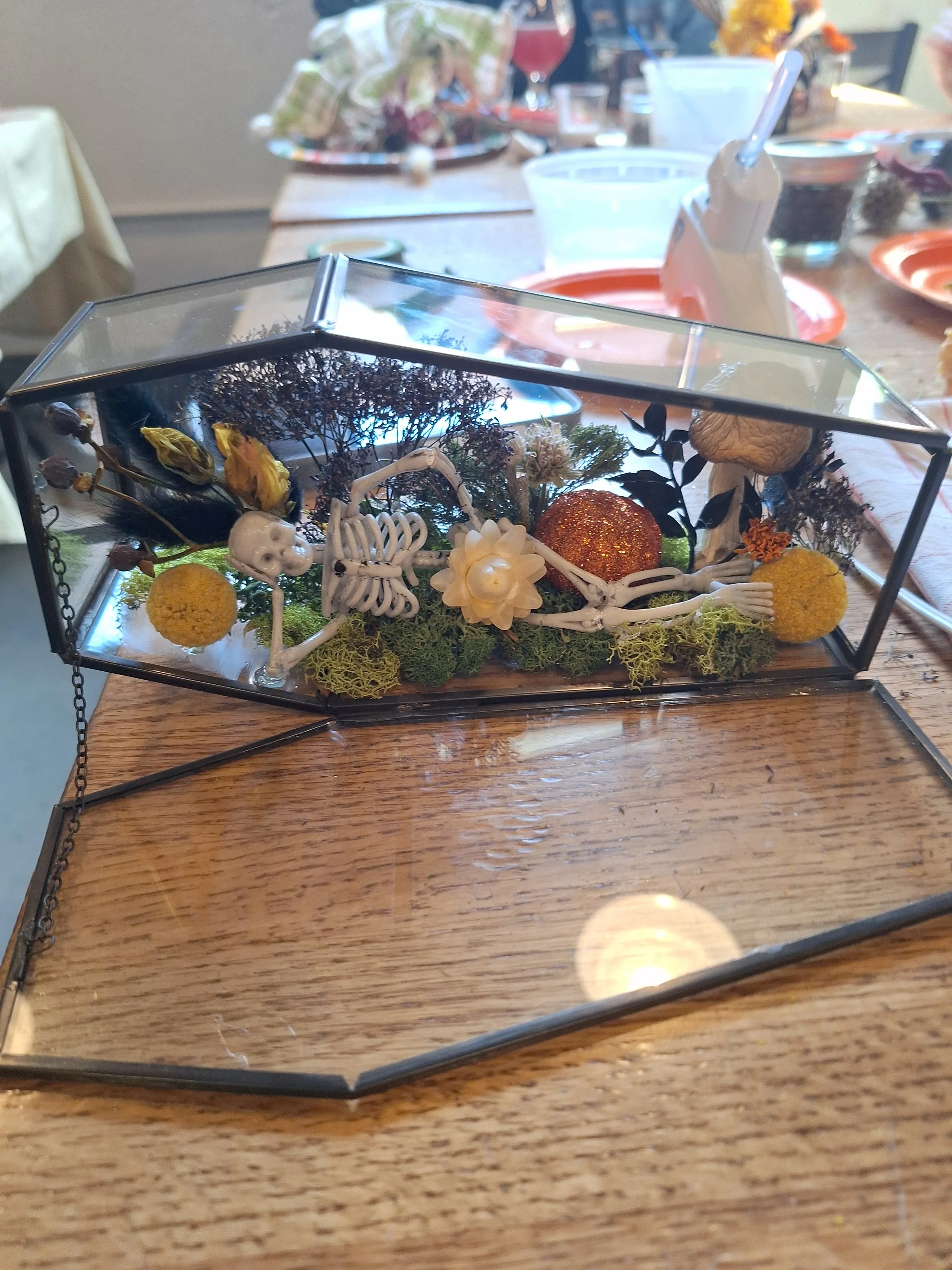A glass box display with a skeleton figure lying among decorative items like flowers, moss, and small objects, on a wooden table.