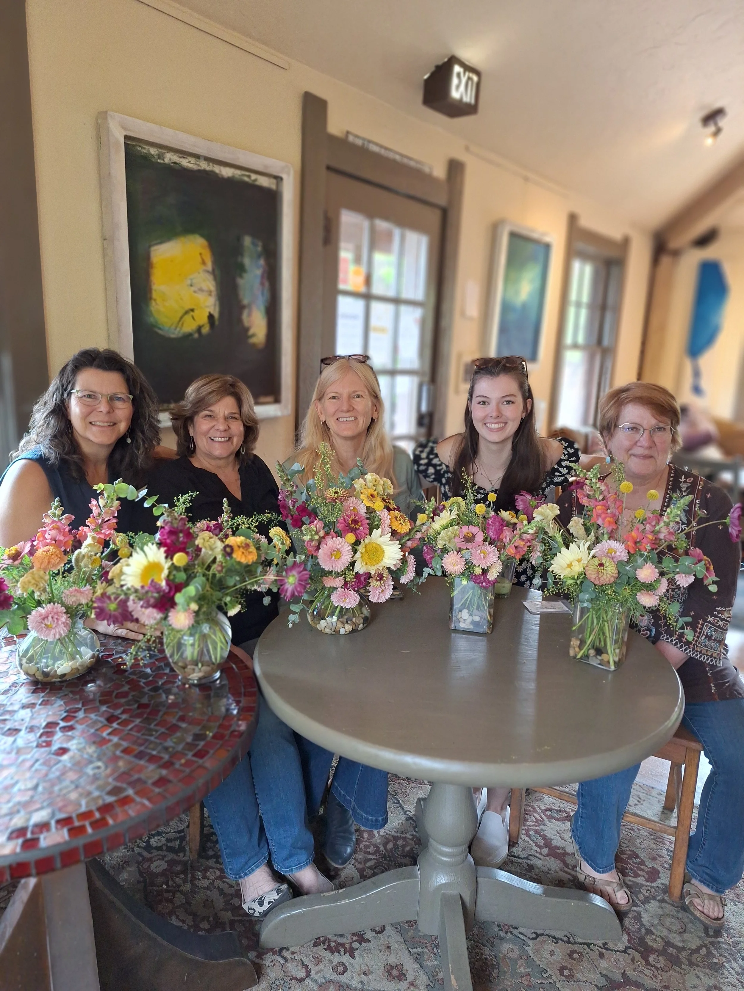 Five women sitting at a table with colorful flower arrangements in glass vases.