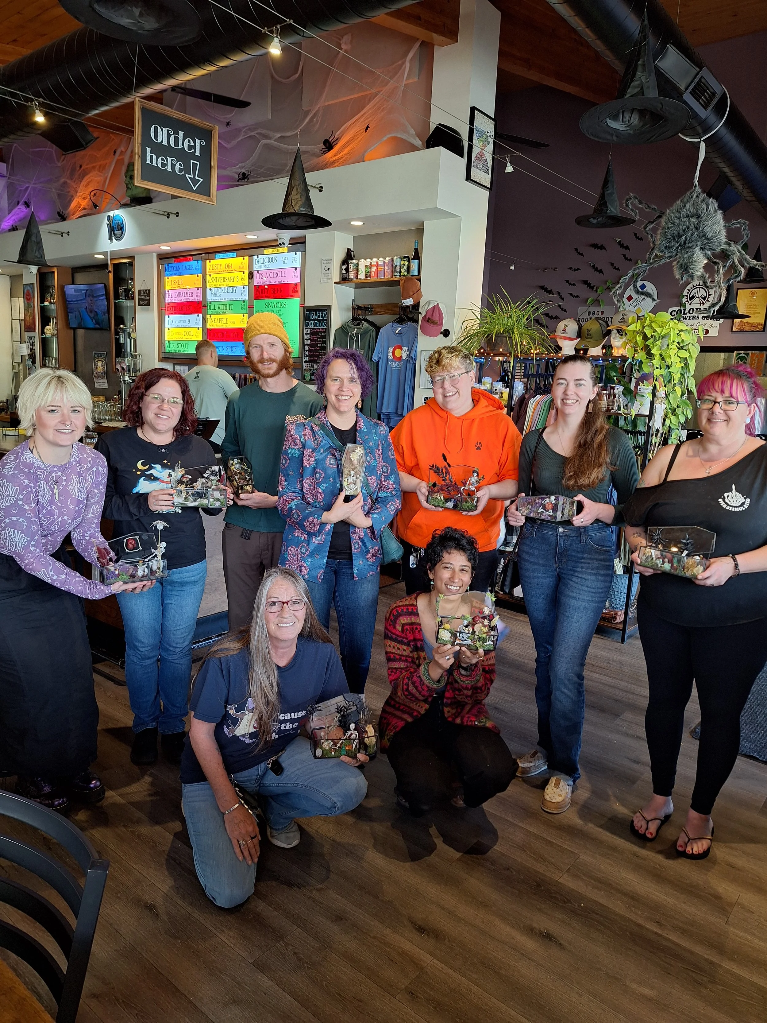 A group of ten people, smiling and holding decorated boxes, gathered in a restaurant or cafe decorated with Halloween-themed decorations, including cobwebs and hanging bats.