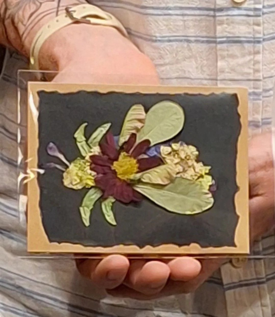 Person holding a framed pressed flower art with a large central flower and surrounding leaves on black background.