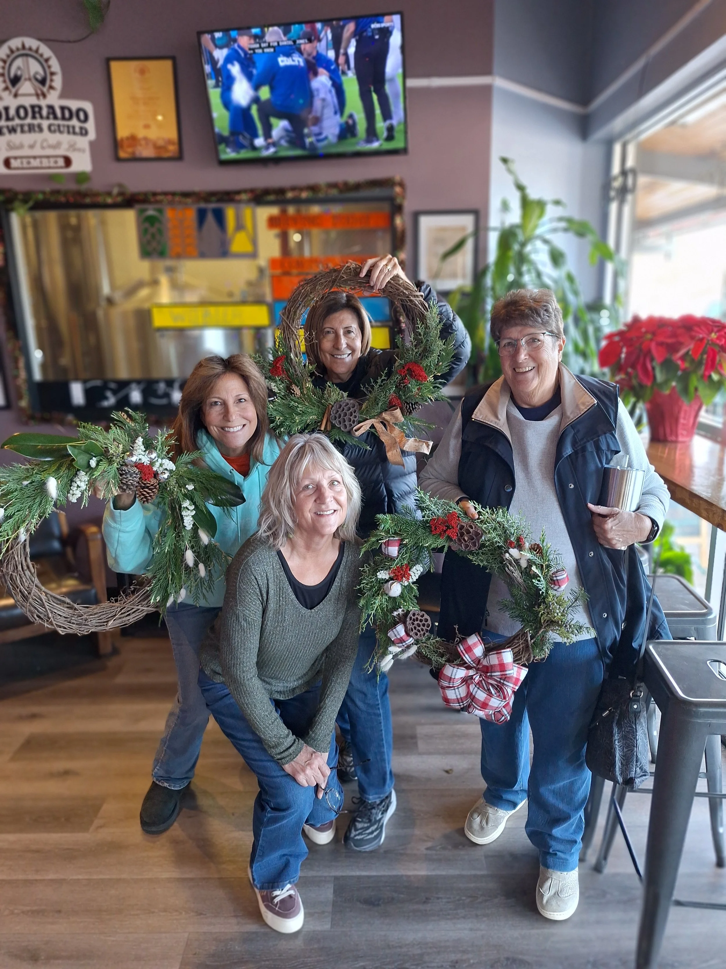Four women smiling and holding Christmas wreaths inside a decorated room with a TV, plants, and poinsettia plants.
