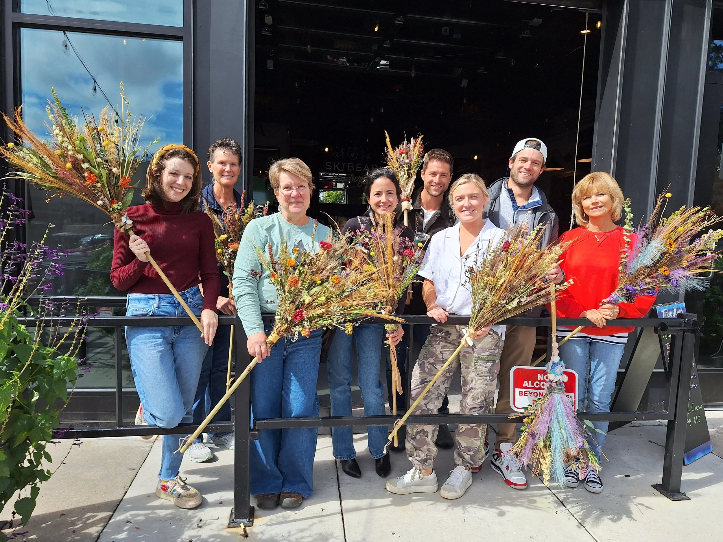 Group of nine people standing outside a building, holding colorful dried flower arrangements, smiling for the photo.