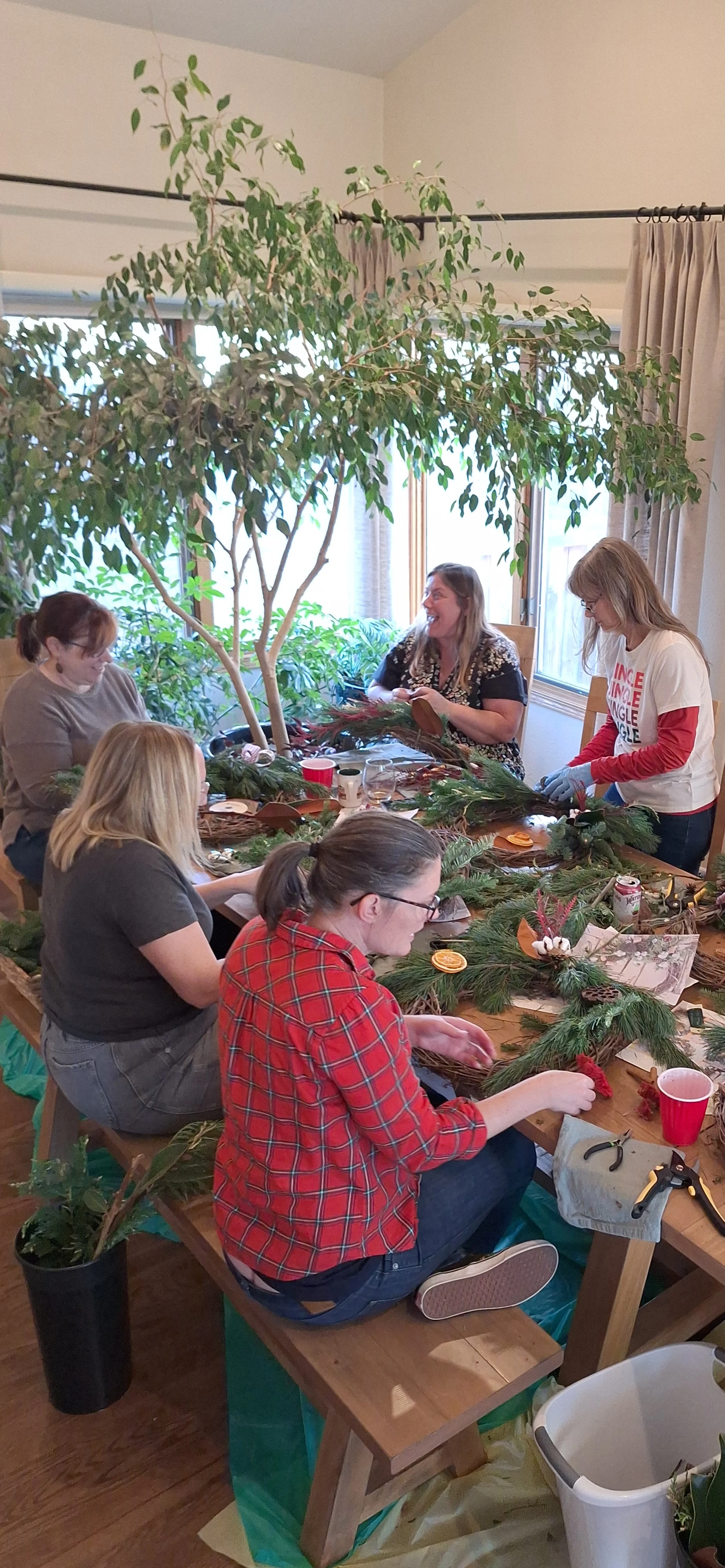 A group of people sitting at a long table making holiday wreaths and arrangements with greenery, pine, and festive decorations in a bright, cozy room with large windows and potted plants.
