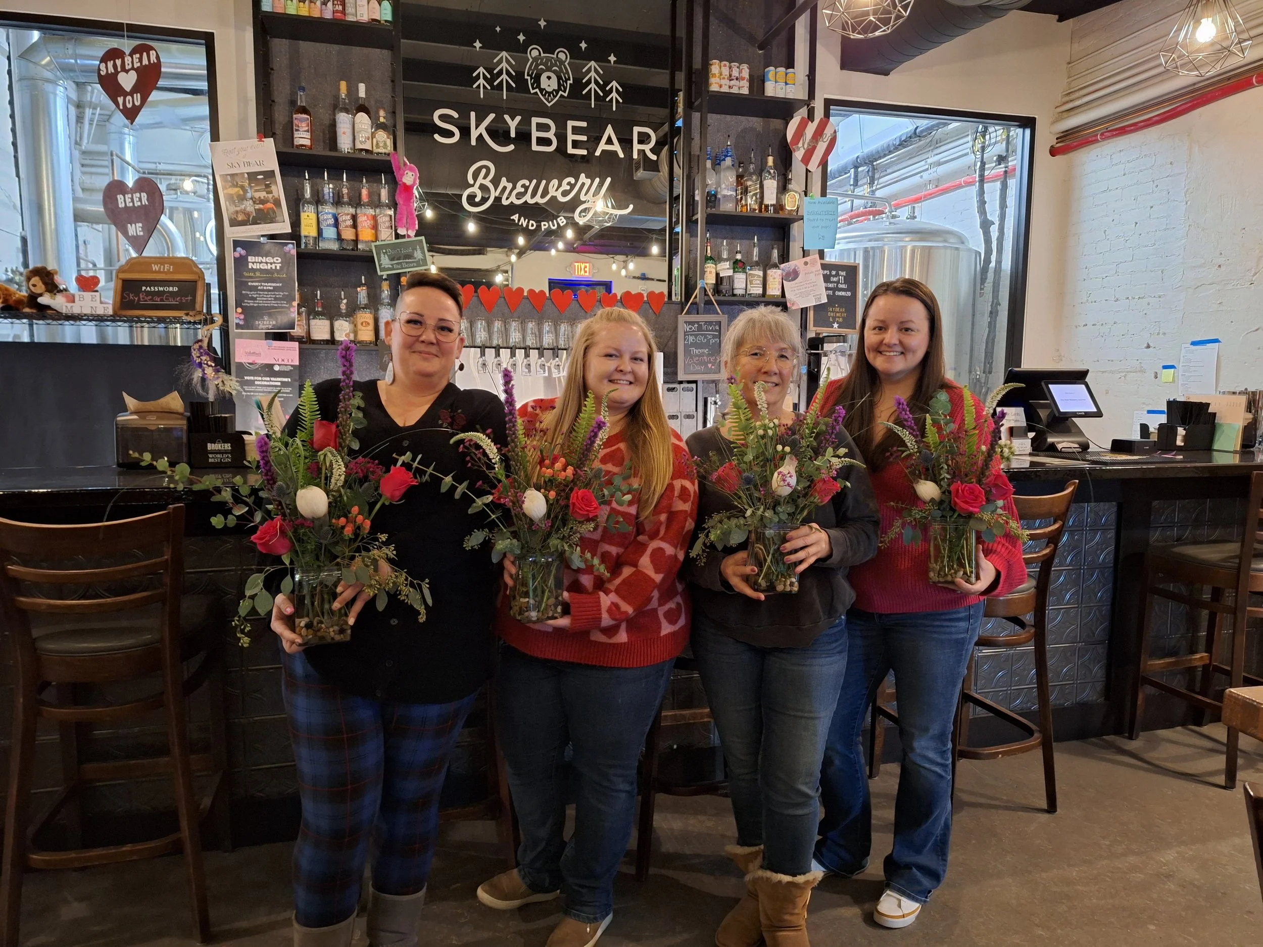 Four women in a brewery, holding floral arrangements in glass vases, standing in front of a bar with bottles on shelves and a large sign that reads "SkyBear Brewery."