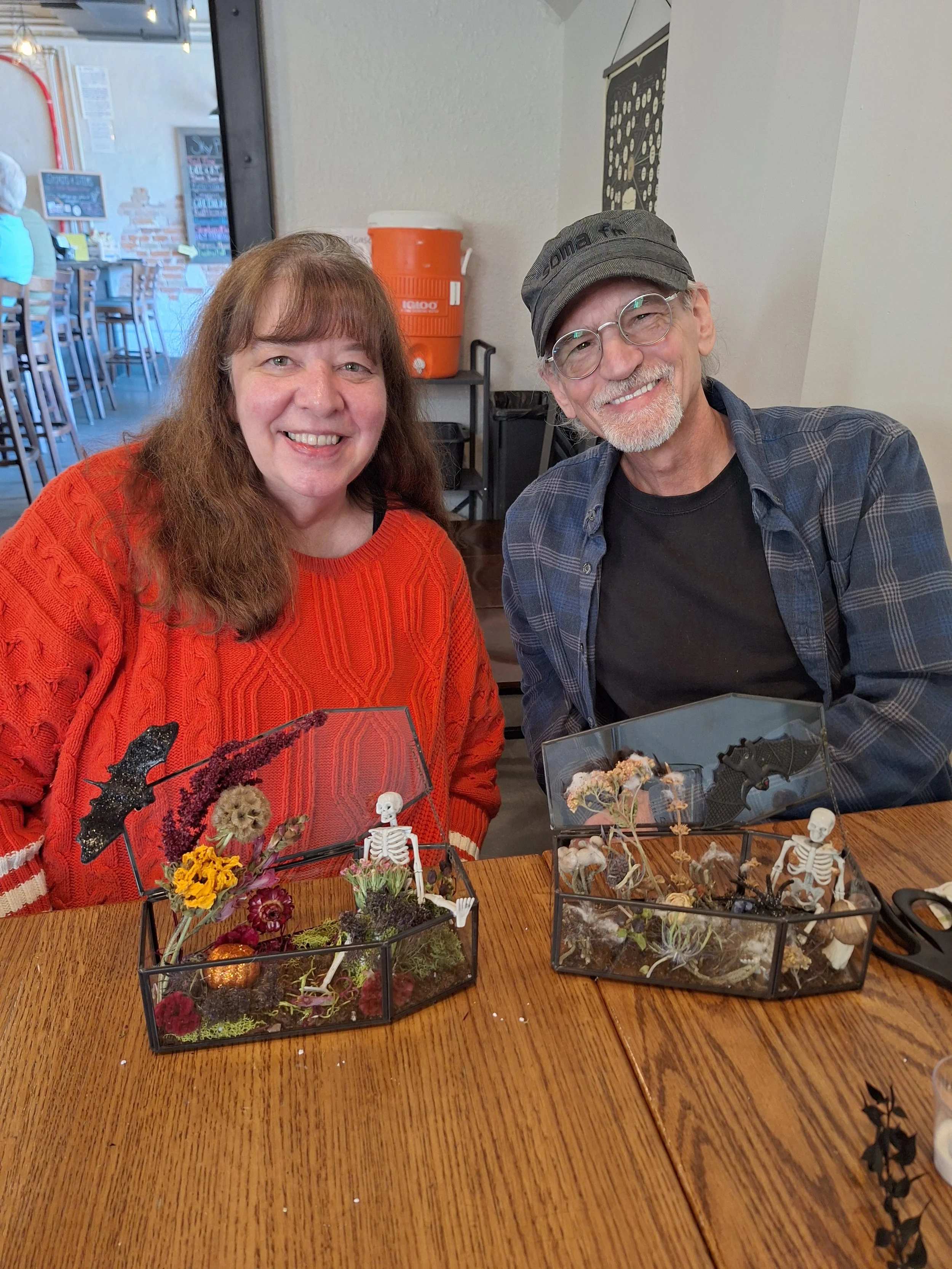 Two smiling people, a woman in an orange sweater and a man in a gray hat and glasses, sitting at a table with Halloween-themed terrariums filled with small skeletons, spiders, and decorative plants.