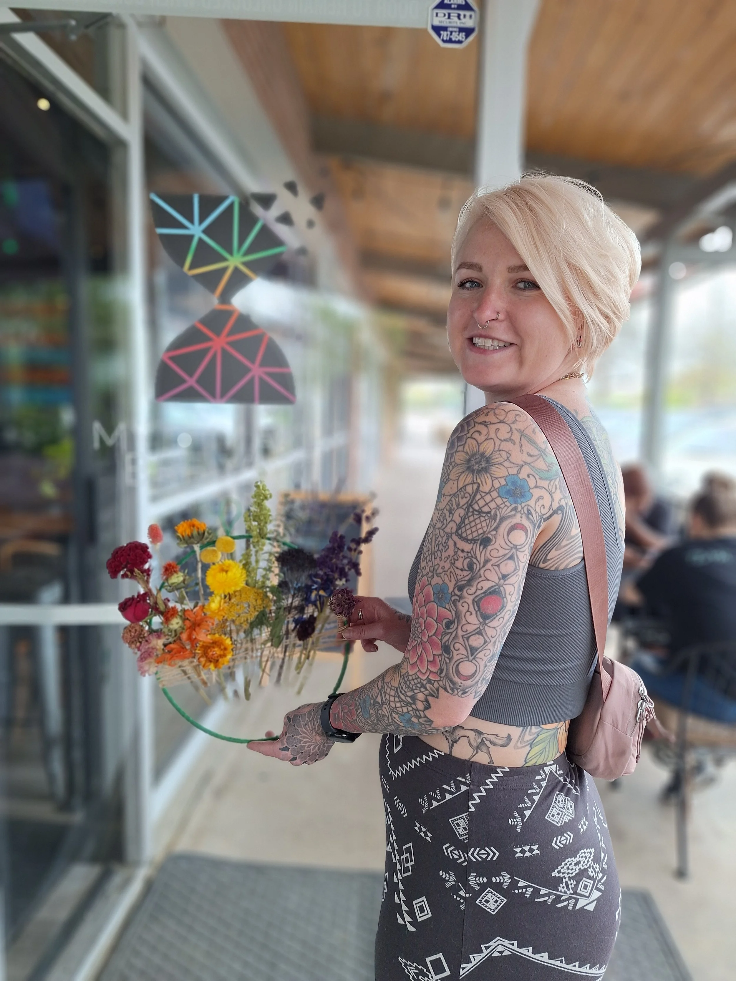 A smiling woman with short blonde hair and tattoos on her arms and torso, holding a small basket of colorful flowers, standing outside a storefront with a glass window, a wooden roof, and outdoor seating.