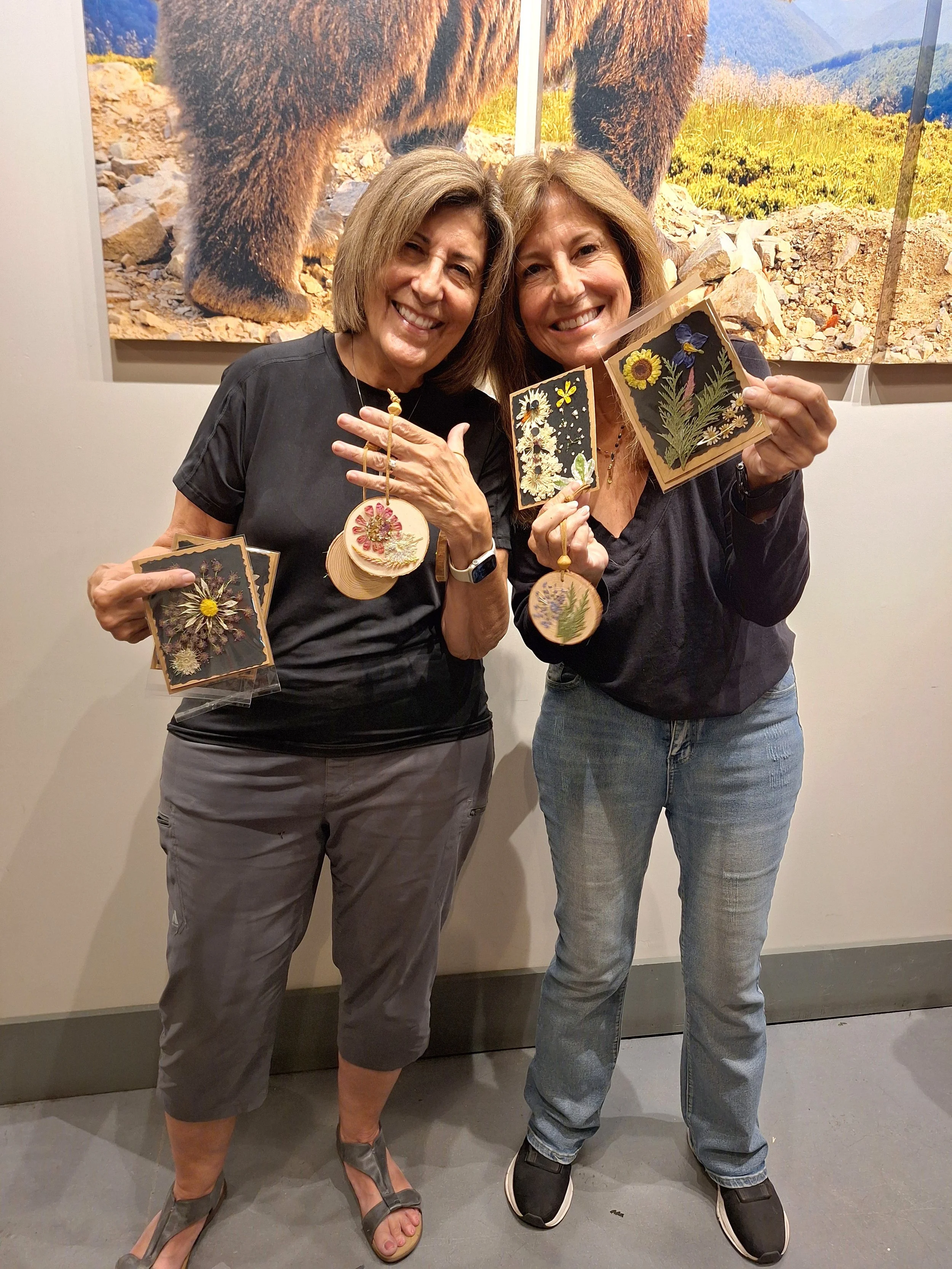 Two women smiling and holding embroidered leaf and flower patches. They are standing in front of a large mountain bear photograph and are holding matching embroidery pieces.