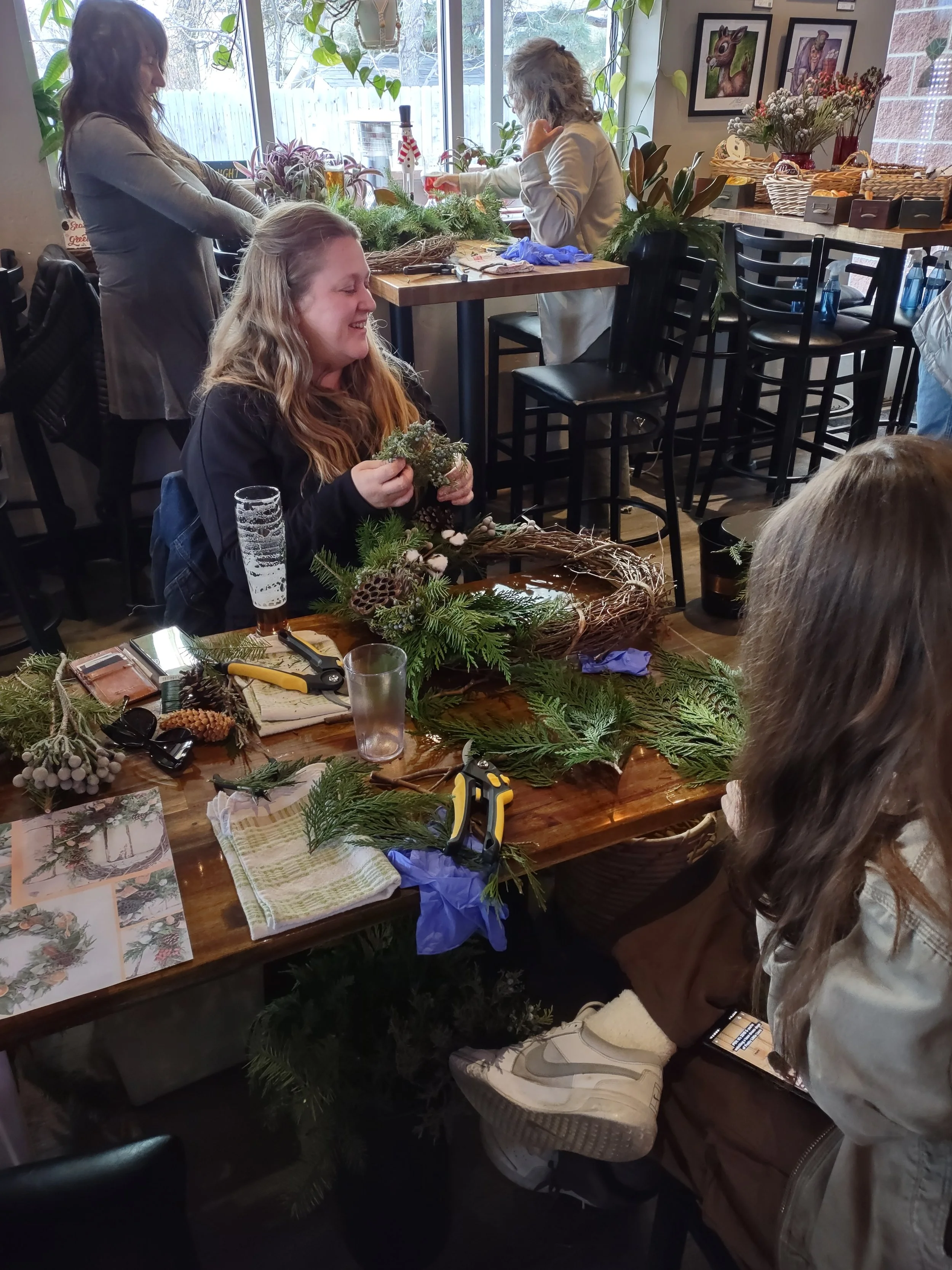 Group of women creating holiday wreaths and decorations at a table inside a cozy room with large windows and framed artwork on the wall.