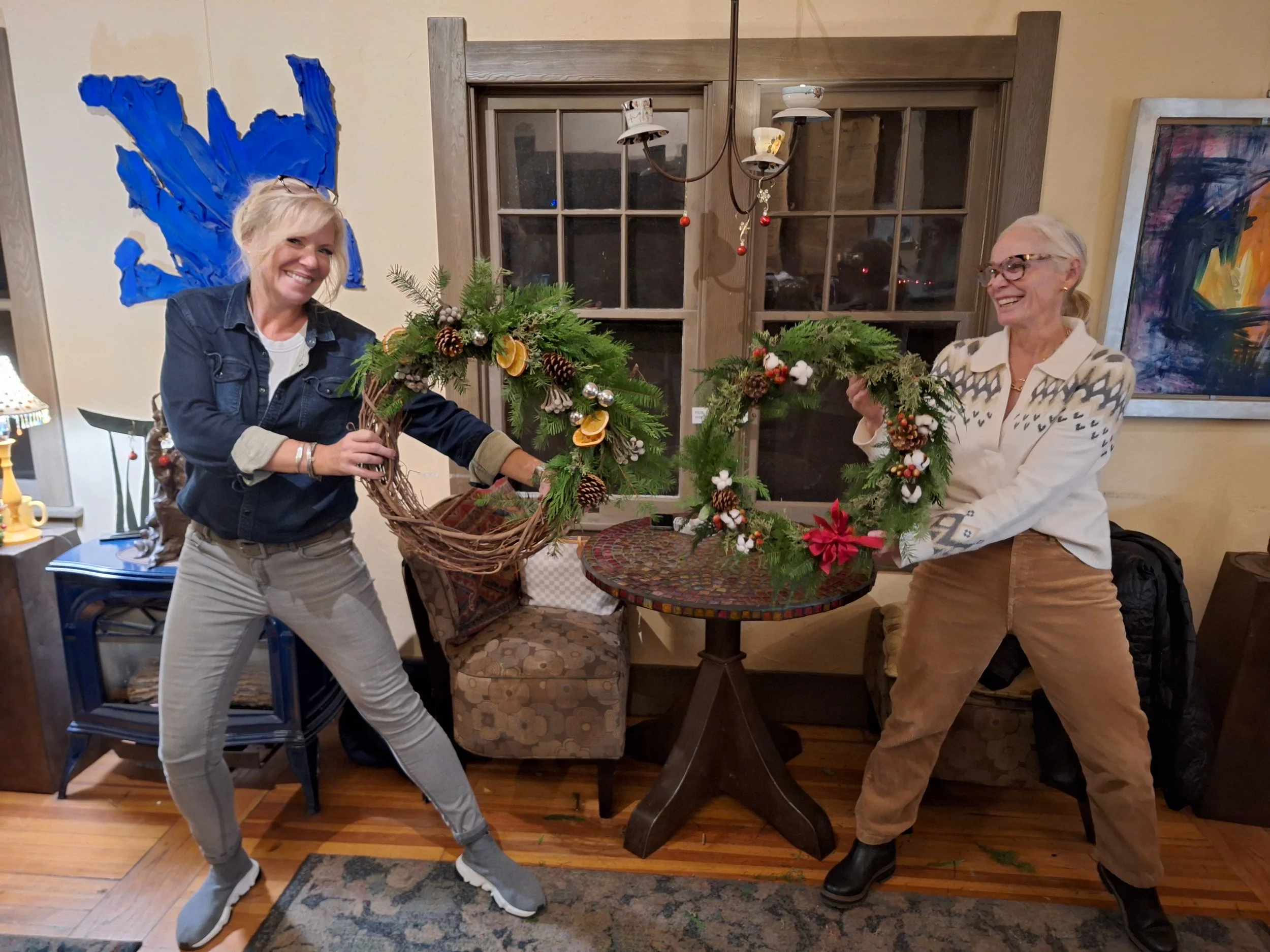 Two women smiling and holding Christmas wreaths inside a cozy room with wooden floors, a round table, and colorful artwork on the walls.