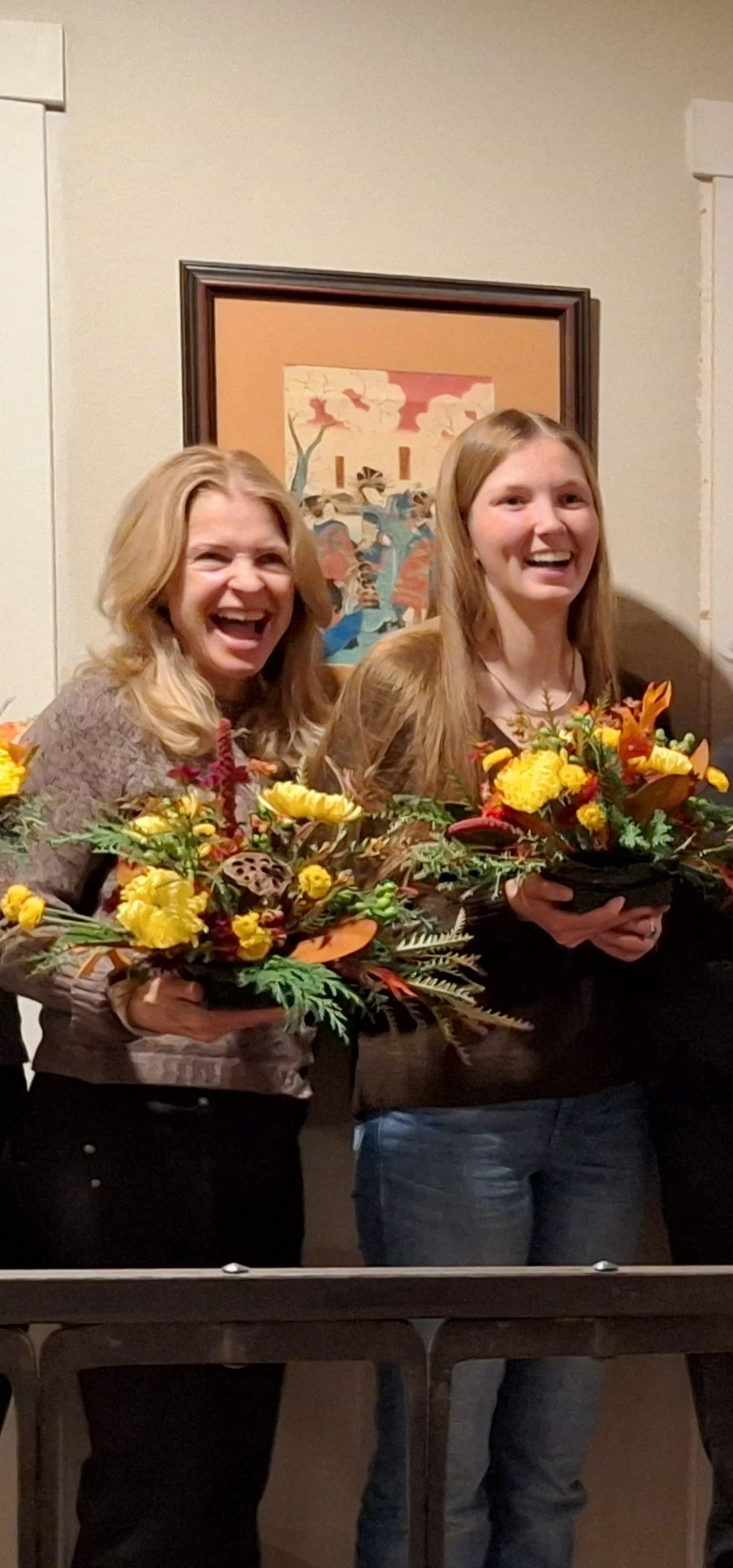 Two women smiling and holding fall flower arrangements, standing indoors in front of a framed artwork.
