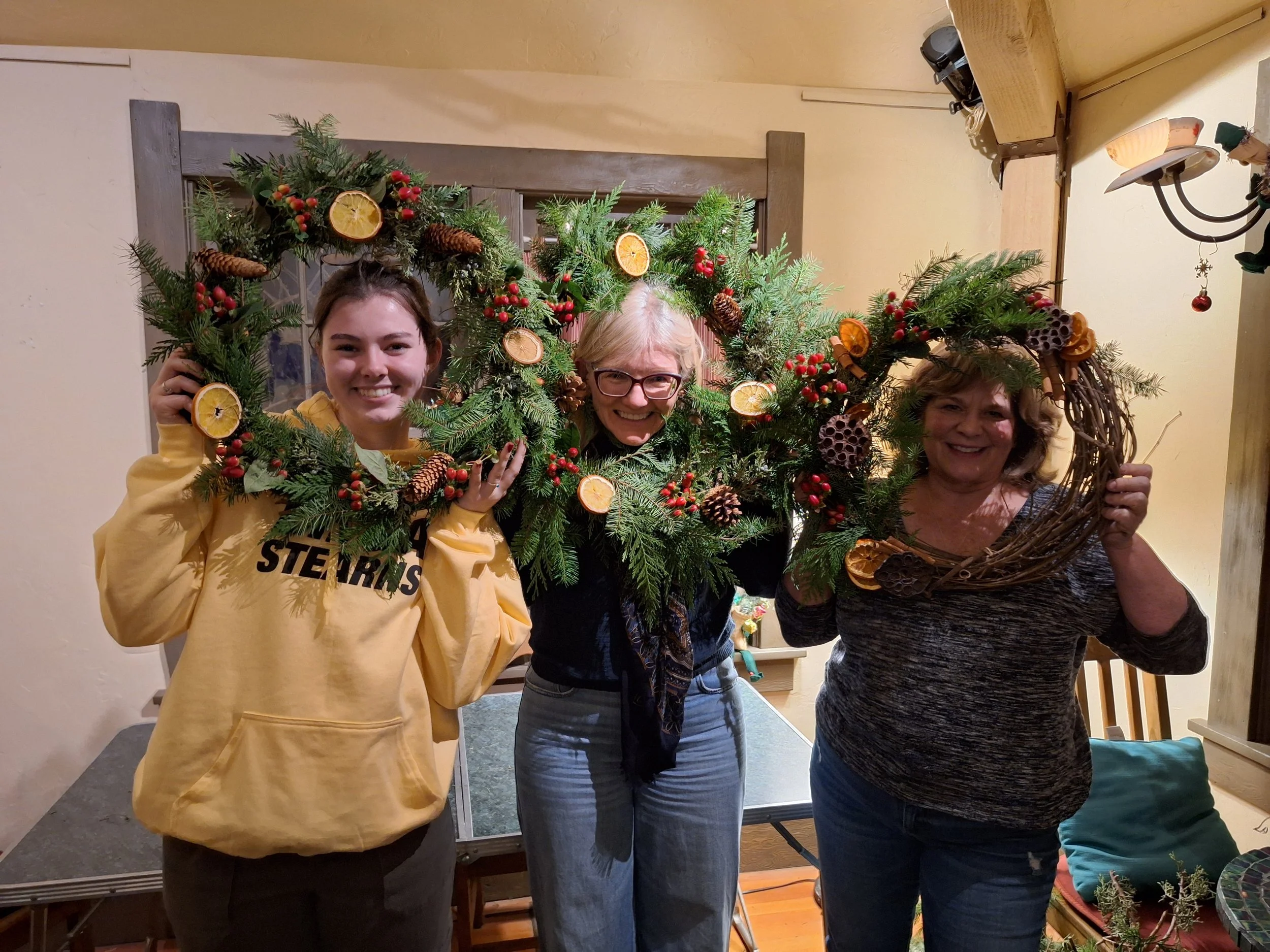 Three women smiling and holding decorated Christmas wreaths made of pine branches with dried orange slices, pinecones, and red berries inside a room