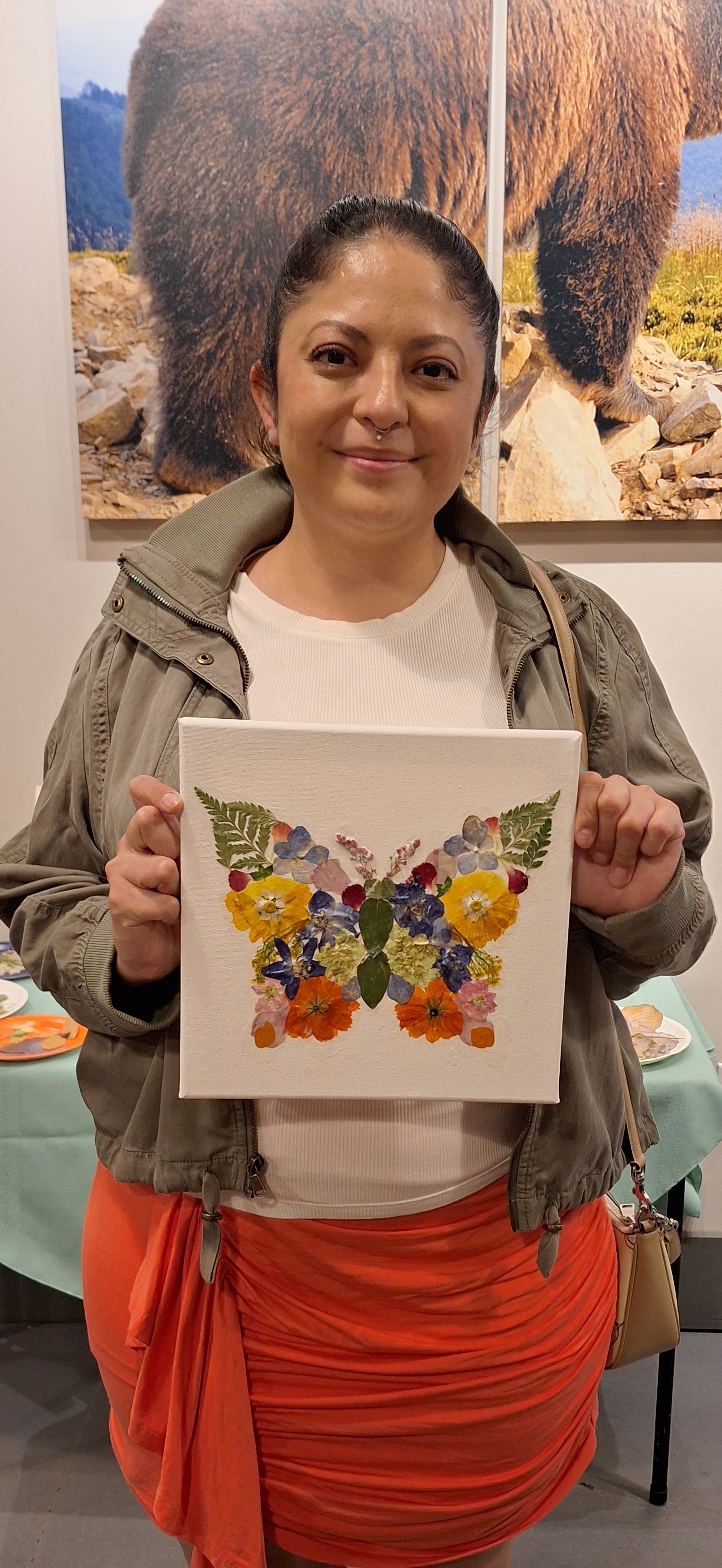 A woman holding a canvas with a butterfly made of pressed flowers, standing in front of a mountain bear photo at an art exhibit.
