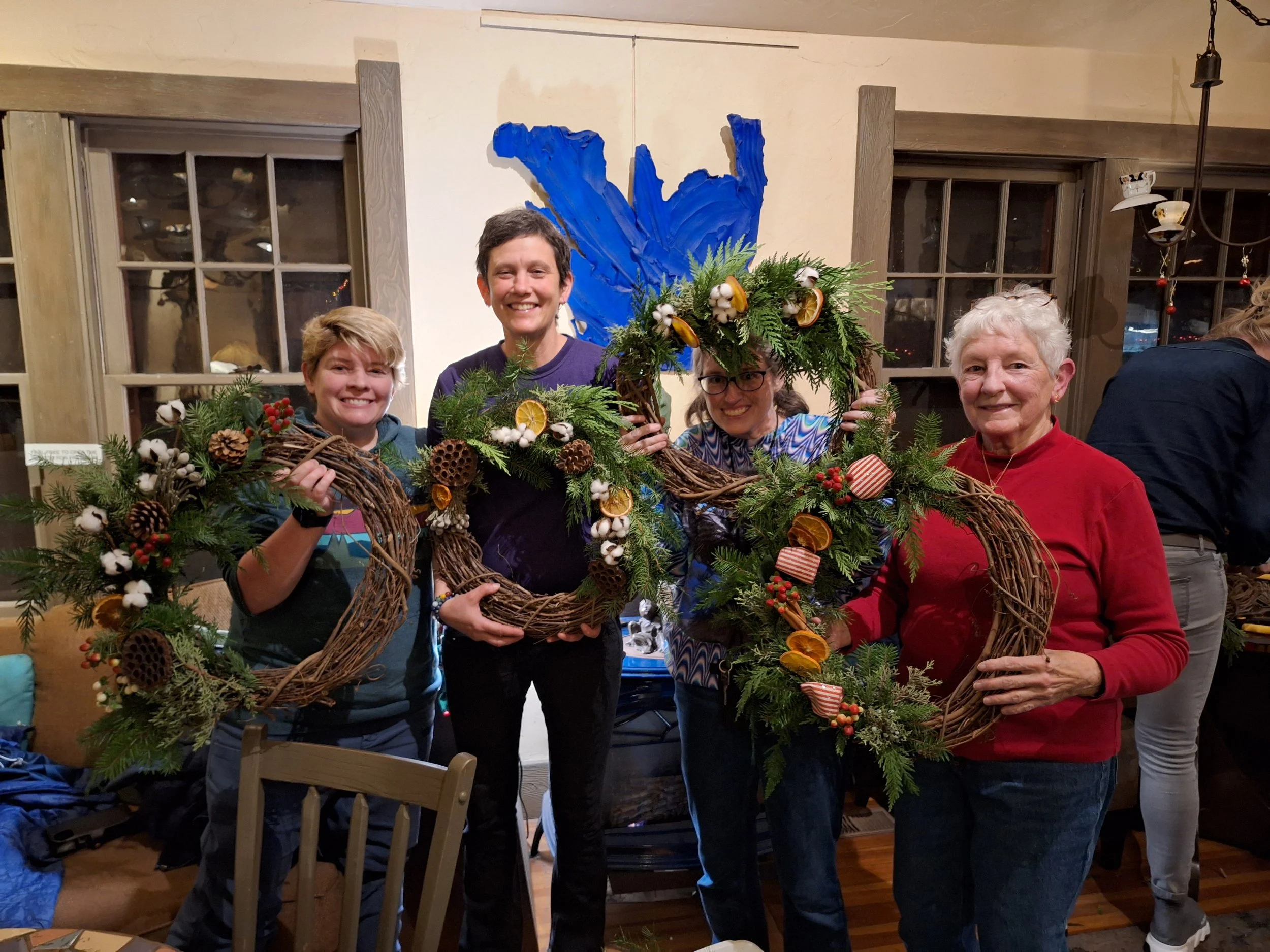 Four women smiling and holding holiday wreaths made of pine, cotton, pine cones, dried fruit, and small American flags, in a cozy indoor setting with wooden window frames and a blue abstract wall art piece in the background.