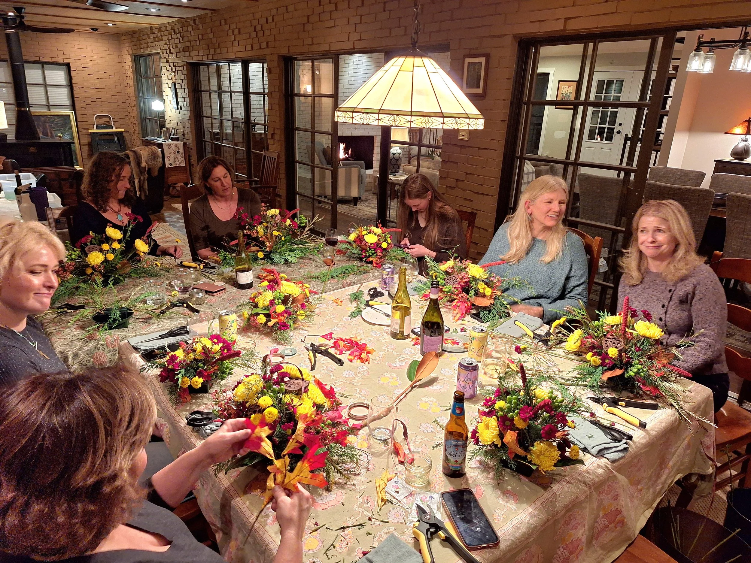 Group of women gathered at a dinner table decorated with floral arrangements, preparing fall-themed centerpieces with leaves and flowers in a cozy home with brick walls and large windows.