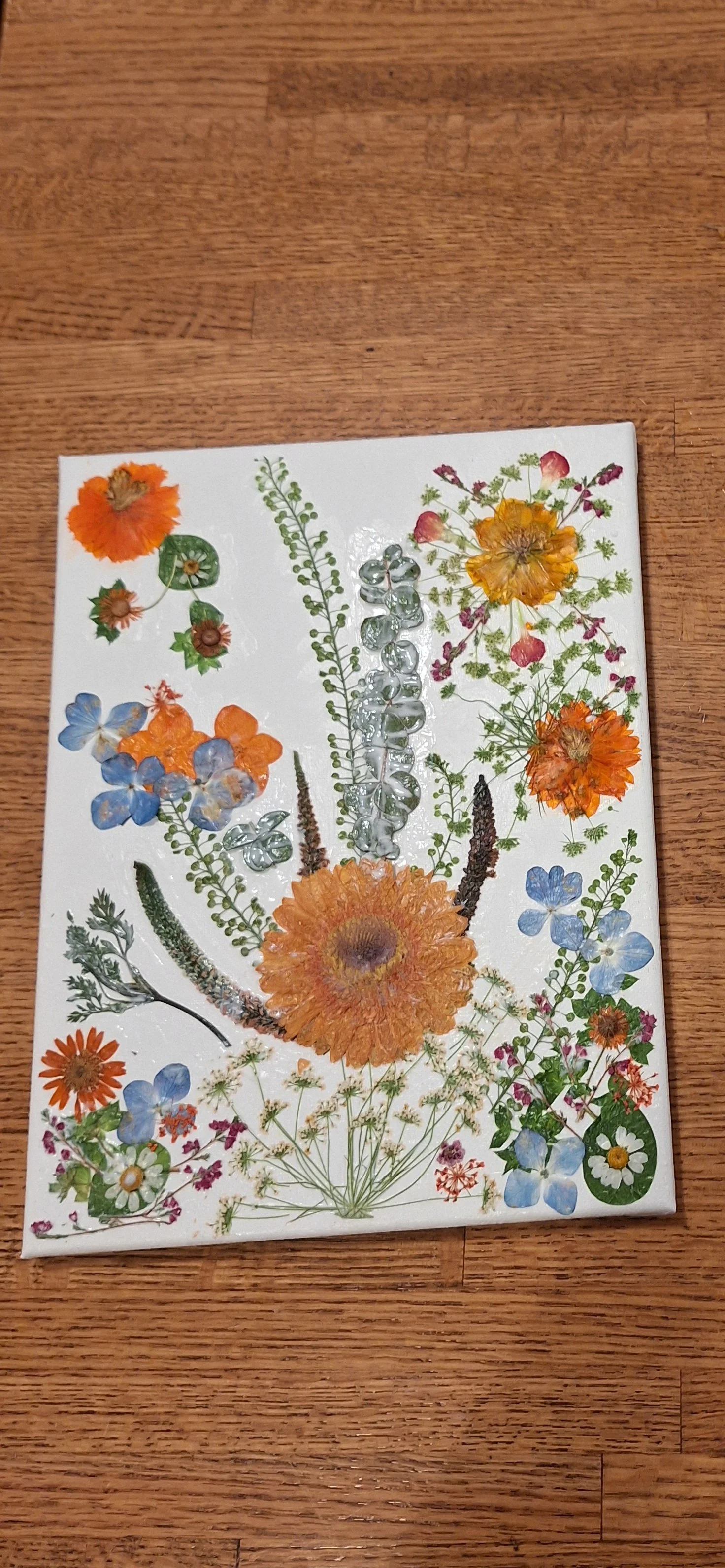 A decorative canvas featuring pressed and dried flowers arranged in a floral design, placed on a wooden surface.