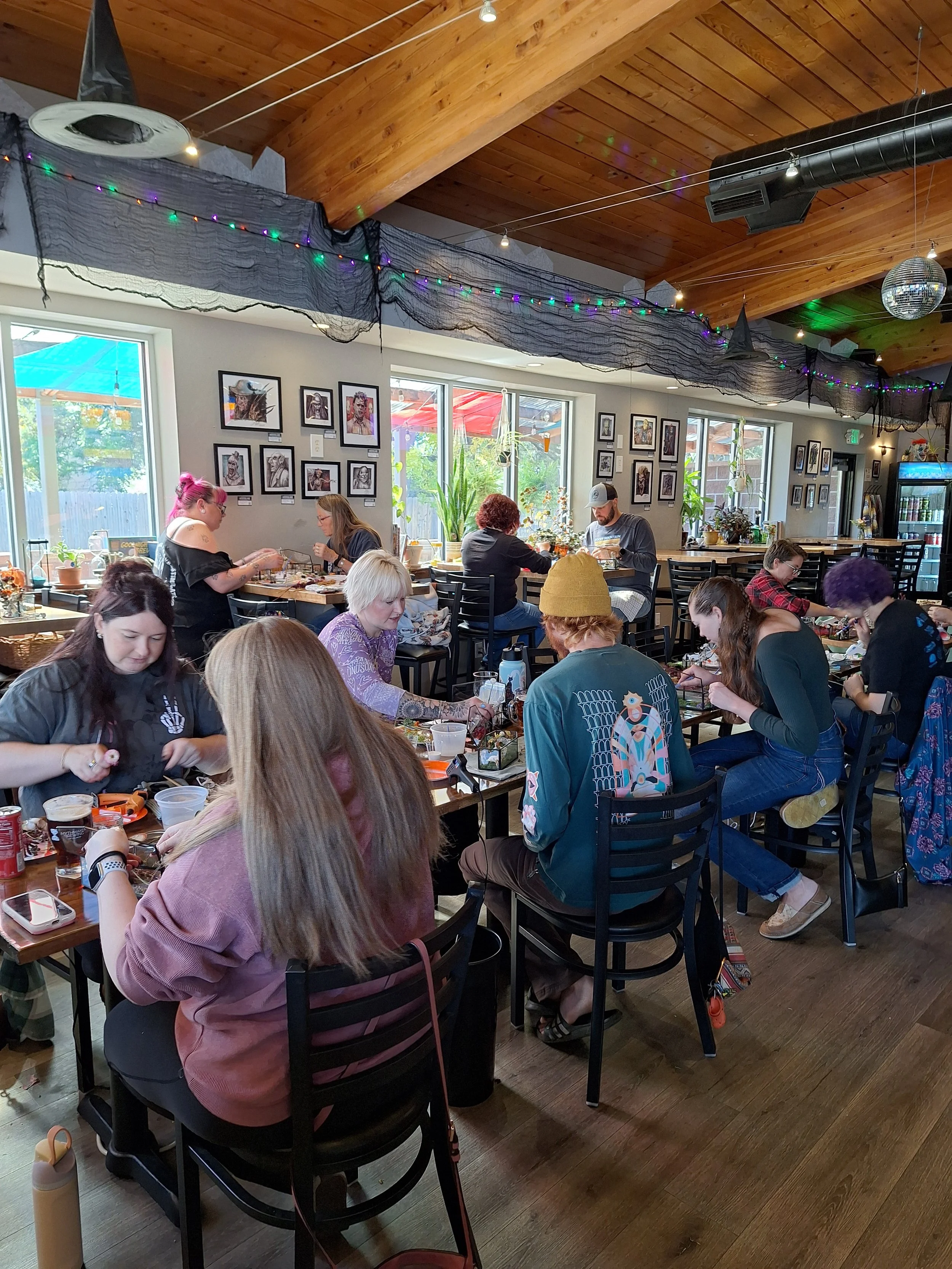 A lively indoor cafe filled with people sitting at tables, some working on crafts or food, with decorated walls, string lights, potted plants, and a wooden ceiling.