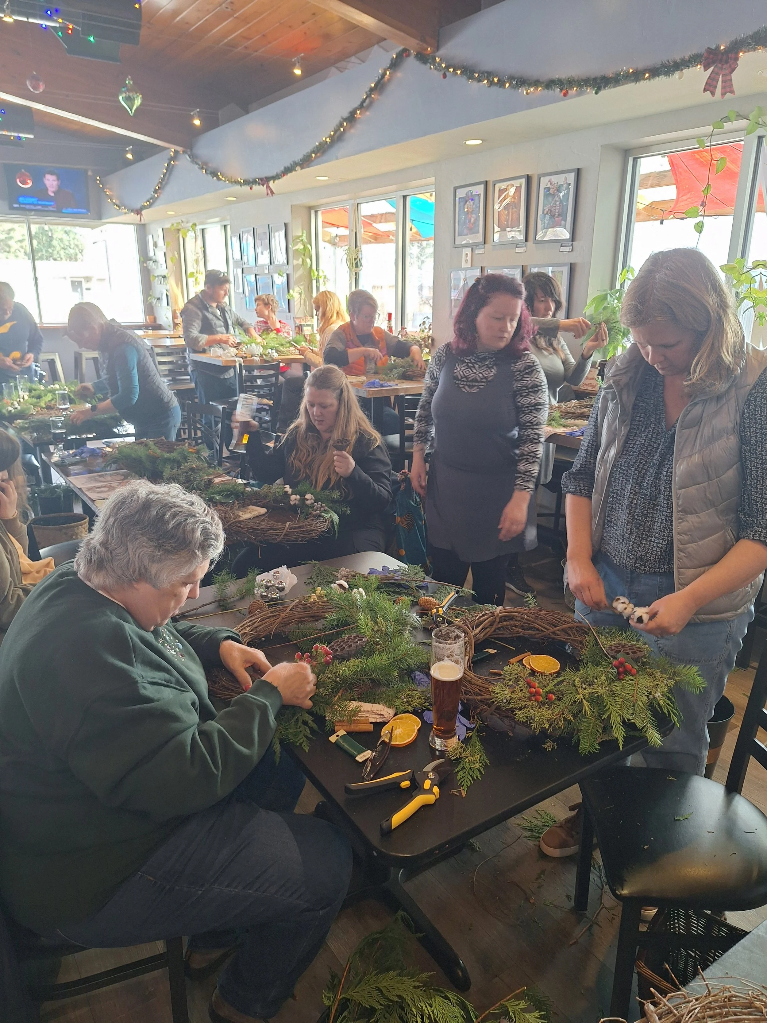 People gathered around tables decorating Christmas wreaths with ornaments, pine branches, and berries in a festive, well-lit room with holiday decorations.