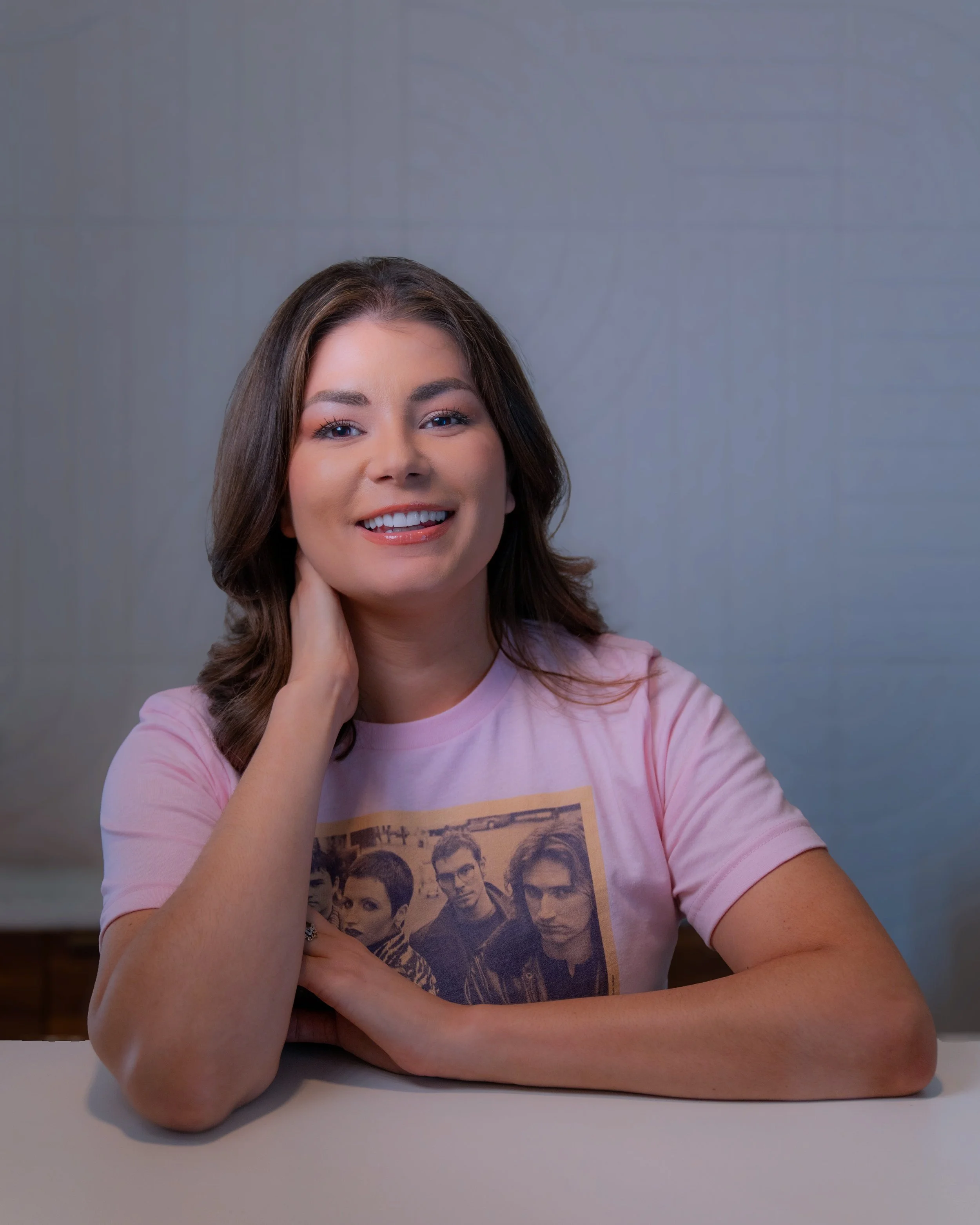 Michelle with shoulder-length brown hair, smiling, wearing a pink T-shirt with a print of a band, sitting at a white table against a plain gray background.