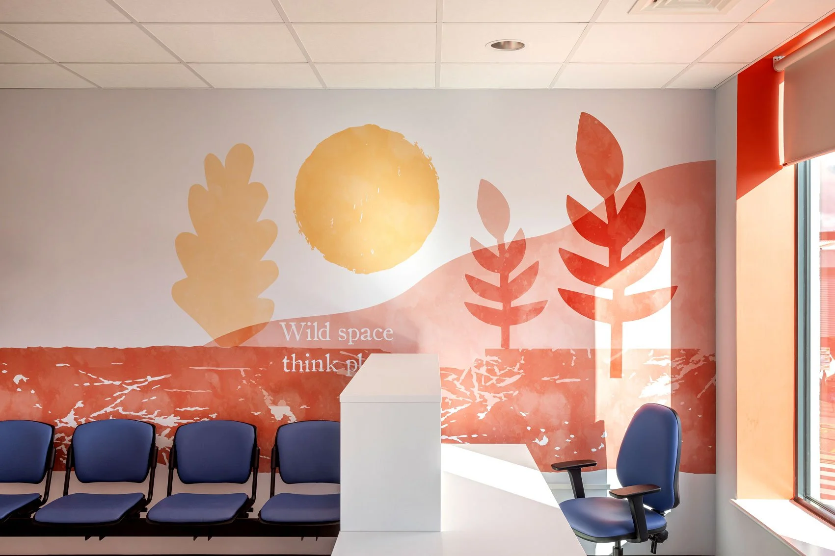 Interior of a room with a wall mural of large abstract plant shapes and a sun, with text that reads 'Wild space think p'. There are five blue chairs in a row, a white counter or podium, and a blue office chair near a window with orange blinds.
