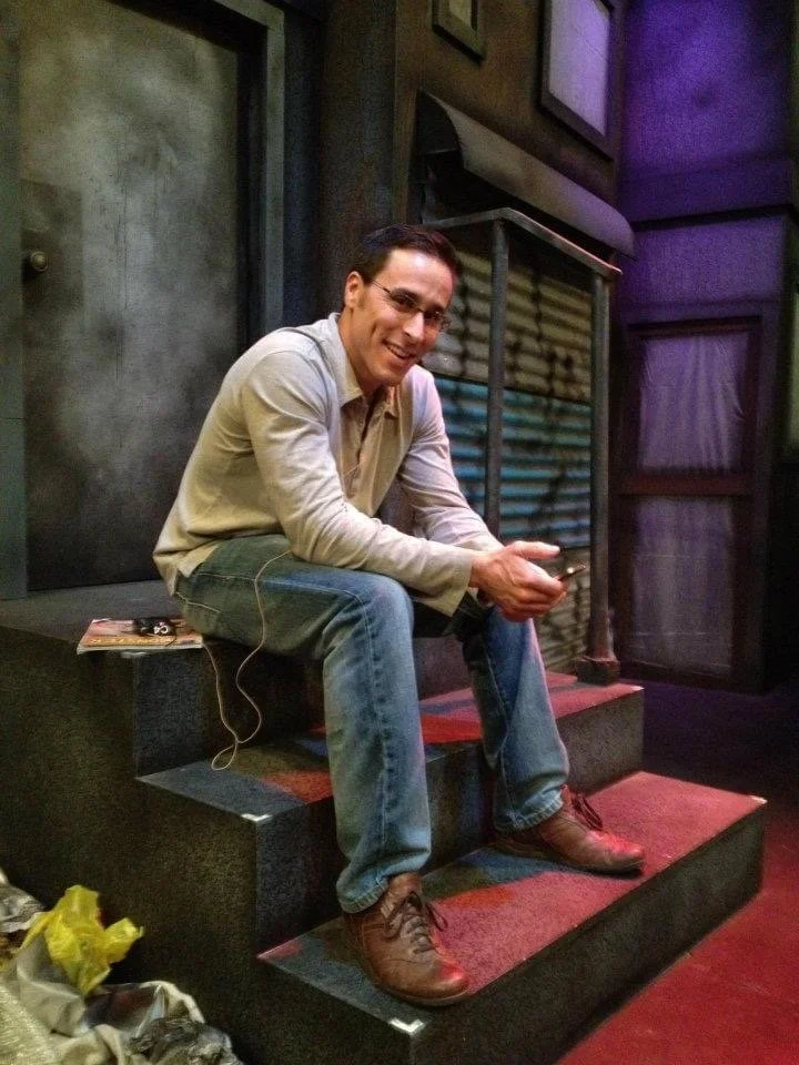 LITTLE SHOP OF HORRORS a comedic musical at Manoa Valley Theatre in Honolulu, Hawaii. Nicholas Gianforti, the Dentist, sitting on stairs in a dimly lit room, smiling and looking at the camera, holding a phone.
