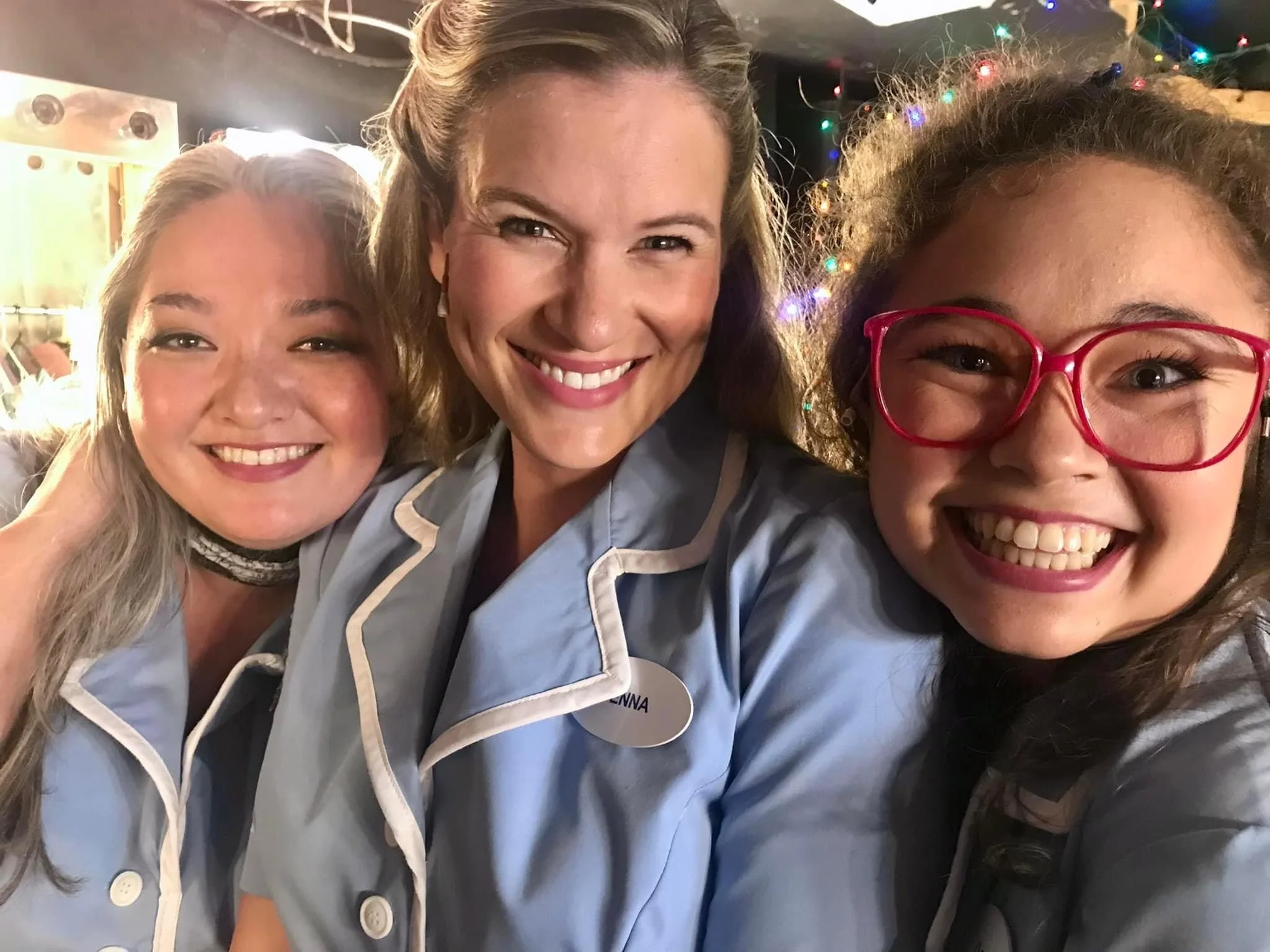 WAITRESS the Broadway musical by Sara Bareilles at Diamond Head Theatre. Three women smiling and taking a selfie together, wearing light blue uniforms, with Christmas lights in the background.