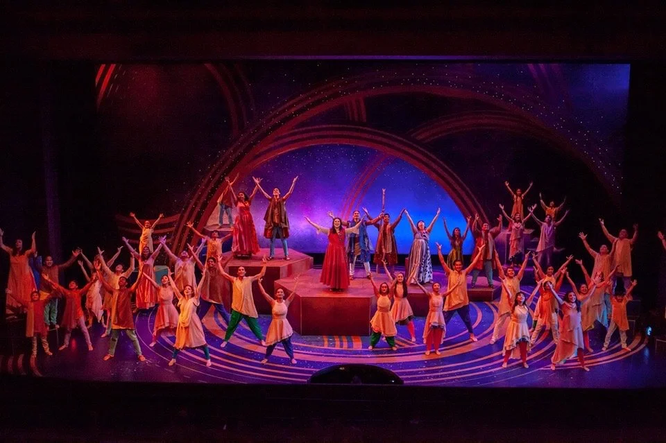 CHILDREN OF EDEN a Broadway musical by I'm A Bright Kid Foundation at Paliku Theatre. A stage performance featuring a large group of dancers in colorful costumes, with some on raised platforms, under vibrant lighting and backdrops.