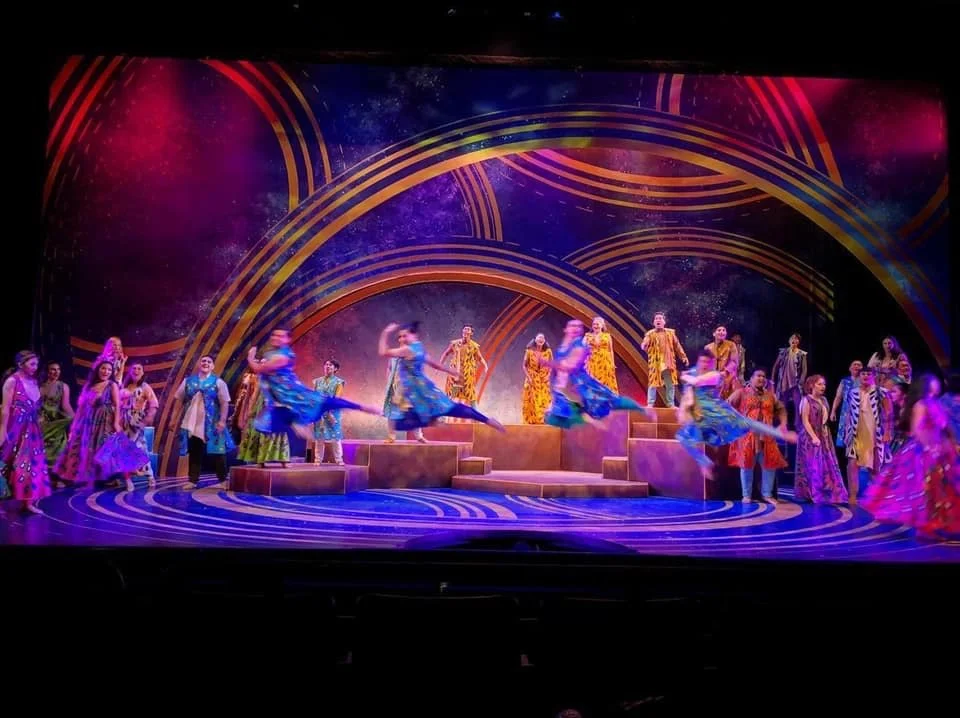 CHILDREN OF EDEN a Broadway musical by I'm A Bright Kid Foundation at Paliku Theatre. A colorful stage performance with dancers in vibrant costumes, some jumping and others standing, set against a cosmic-themed backdrop with swirling patterns.