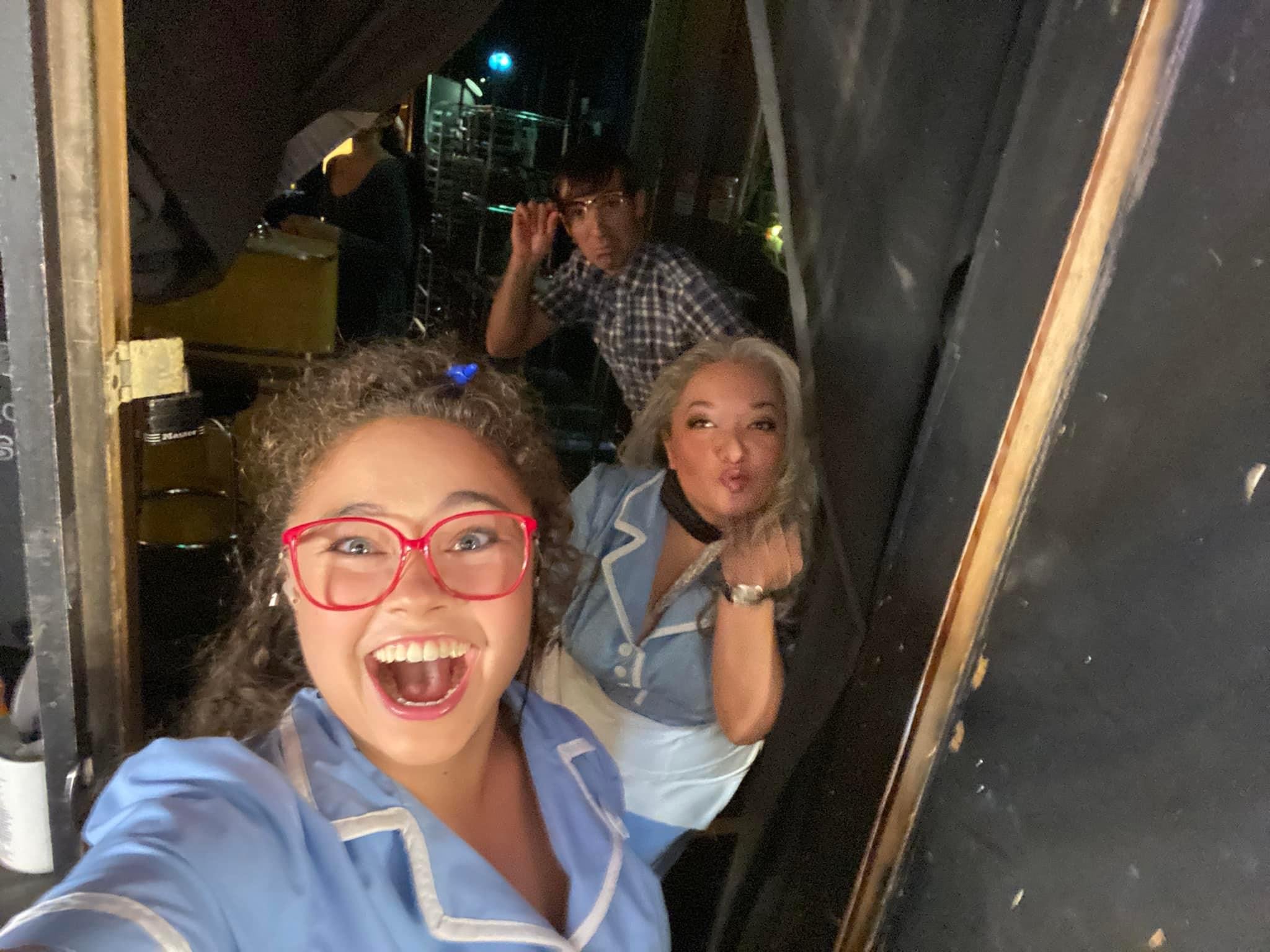 The stars of WAITRESS the Broadway musical. Three people in costumes posing for a selfie backstage at a performance, with one person in the foreground wearing red glasses and an excited expression.