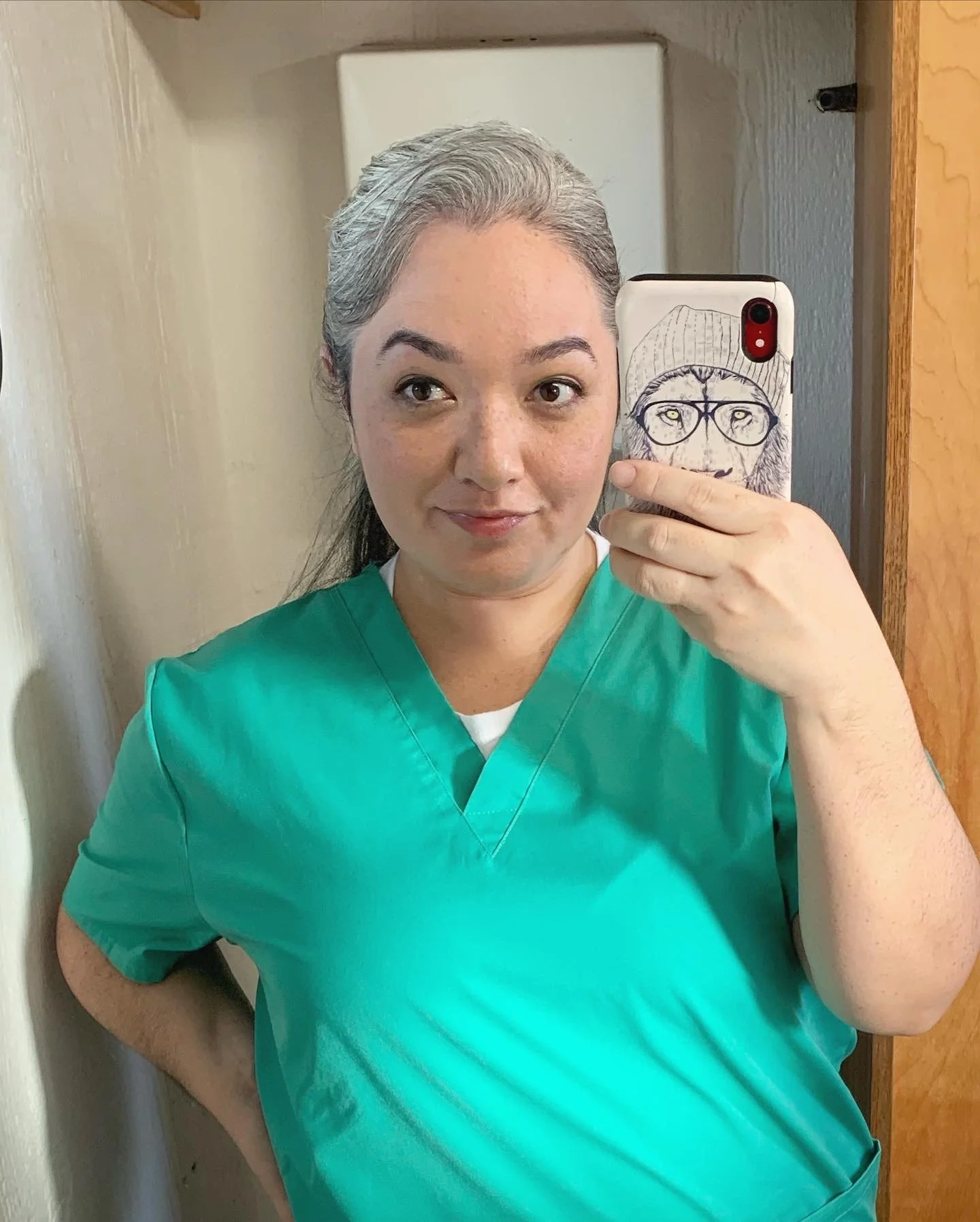 On set filming for MAGNUM P.I. with CBS on the studio lot. Mirror selfie. She is wearing green scrubs and holding a phone with a case featuring a drawing of a face with glasses and a beanie, depicting a lion wearing glasses.