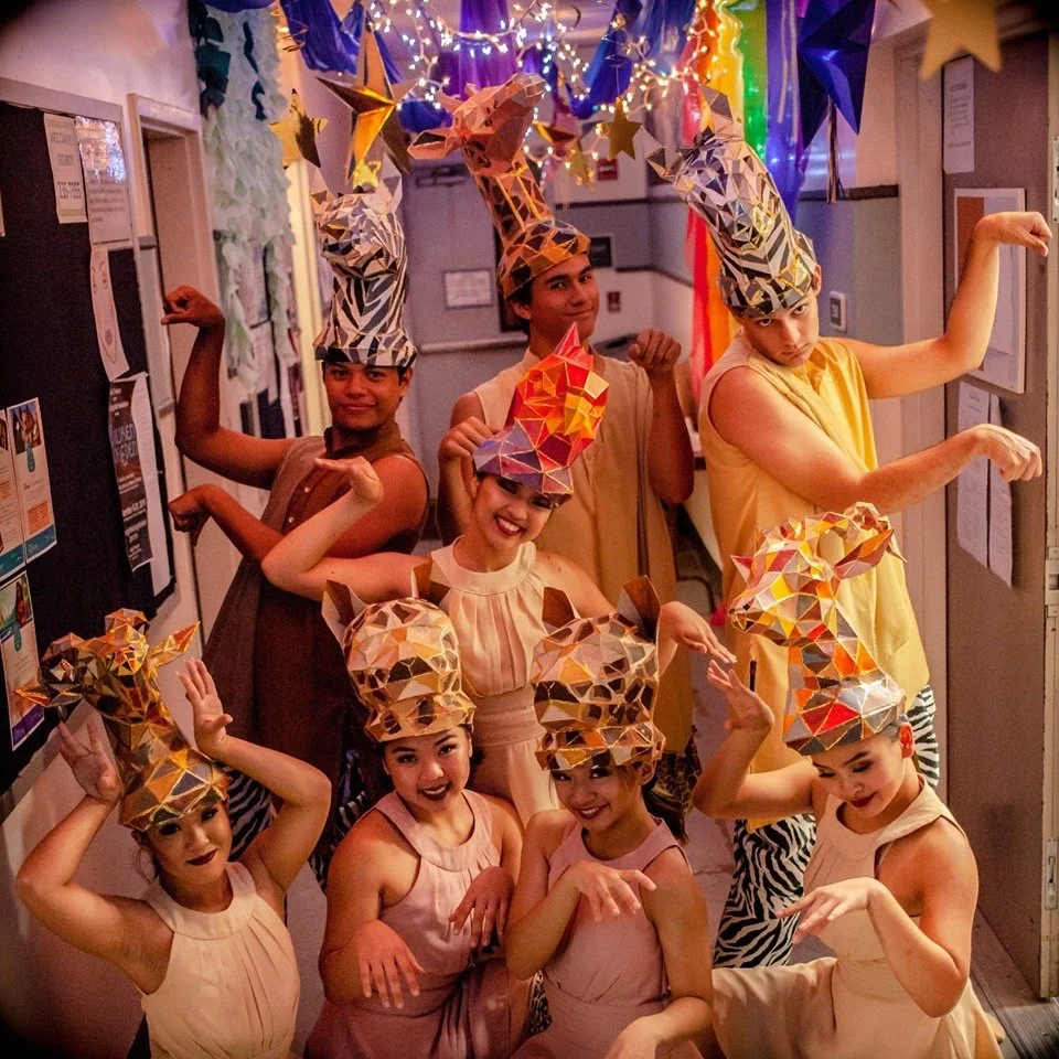 Group of people dressed in colorful costumes with animal-themed hats, striking playful poses in a decorated indoor setting with string lights and colorful hanging decorations.