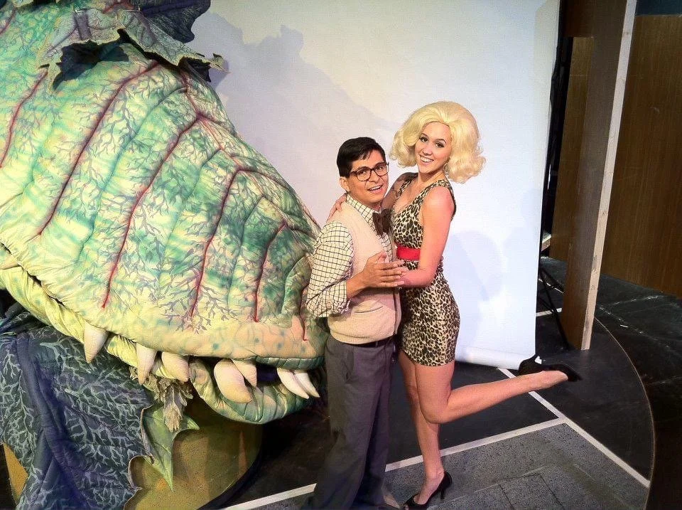 LITTLE SHOP OF HORRORS a comedic musical at Manoa Valley Theatre in Honolulu, Hawaii. Two people in costume posing with a large, dragon-like puppet or prop that has a leafy, textured head and menacing teeth; they are indoors near a white backdrop.