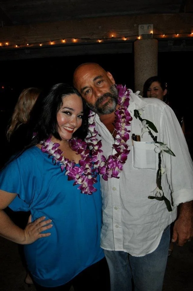 LITTLE SHOP OF HORRORS a comedic musical at Manoa Valley Theatre. A woman with black hair in a blue top and a man with a beard in a white shirt, both wearing purple leis, posing closely together at a social event, with other people in the background.