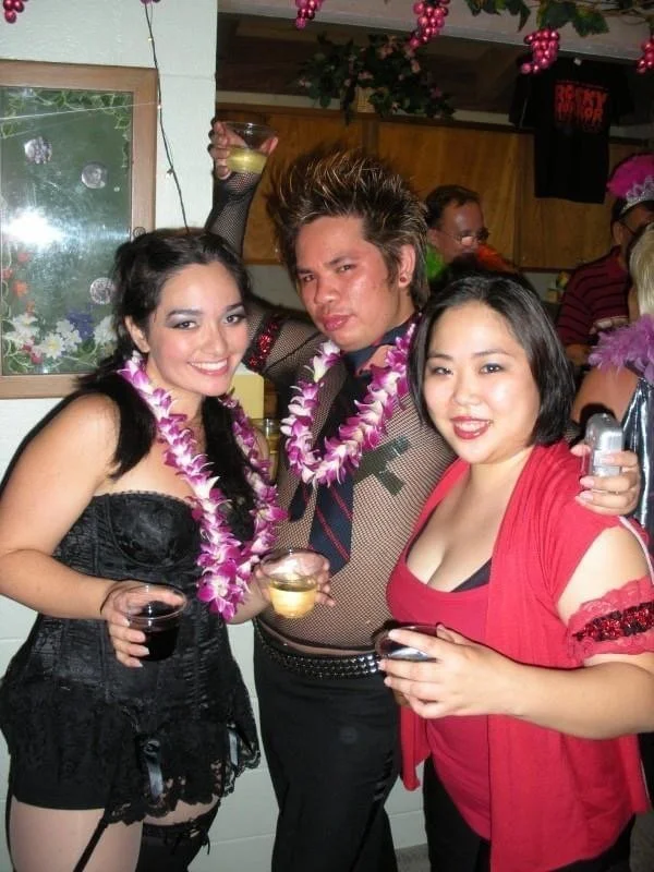 ROCKY HORROR SHOW AT Manoa Valley Theatre. Actress Cassie Favreau-Chung plays Magenta. Group of three people dressed in party attire, wearing leis, holding drinks, and posing together at a festive celebration with pink decorations in the background.