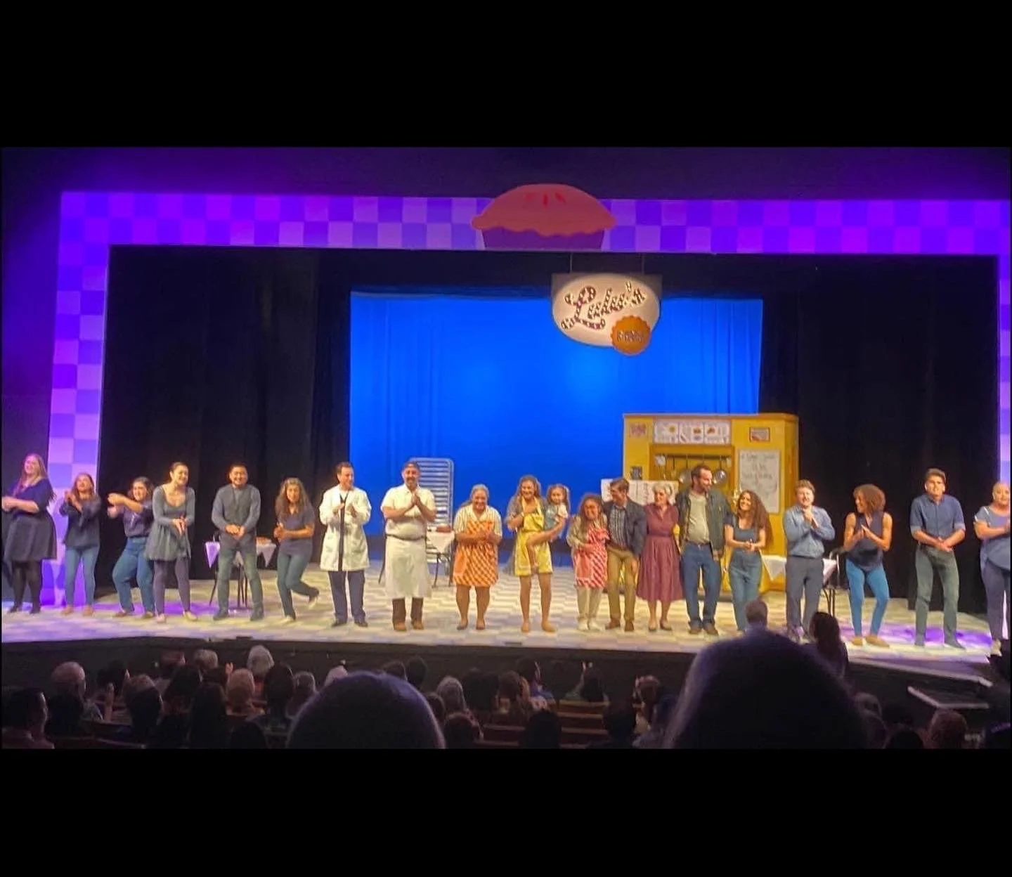 WAITRESS the Broadway musical by Sara Bareilles at Diamond Head Theatre. The cast of a stage play is standing in a line across the stage, clapping and smiling at the audience in a theater.