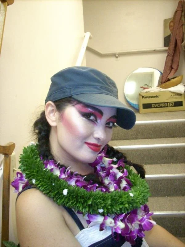 ROCKY HORROR SHOW AT Manoa Valley Theatre. Actress Cassie Favreau-Chung plays Magenta. A woman wearing a dark cap, makeup, and a flower lei, sitting on stairs in a room with a mirror, cardboard box, and a coat hanging on the wall.