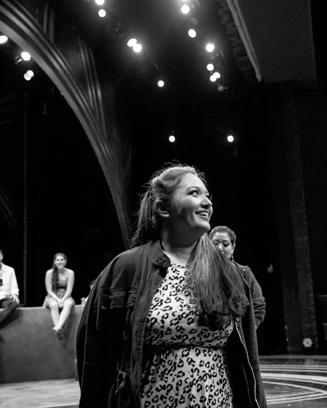 Hawaii actress and singer, Cassie Favreau-Chung, onstage at Paliku Theatre for Children of Eden during vocal warmups. A woman smiling, standing on a stage with a dark background and bright stage lights overhead. I'm A Bright Kid Foundation.