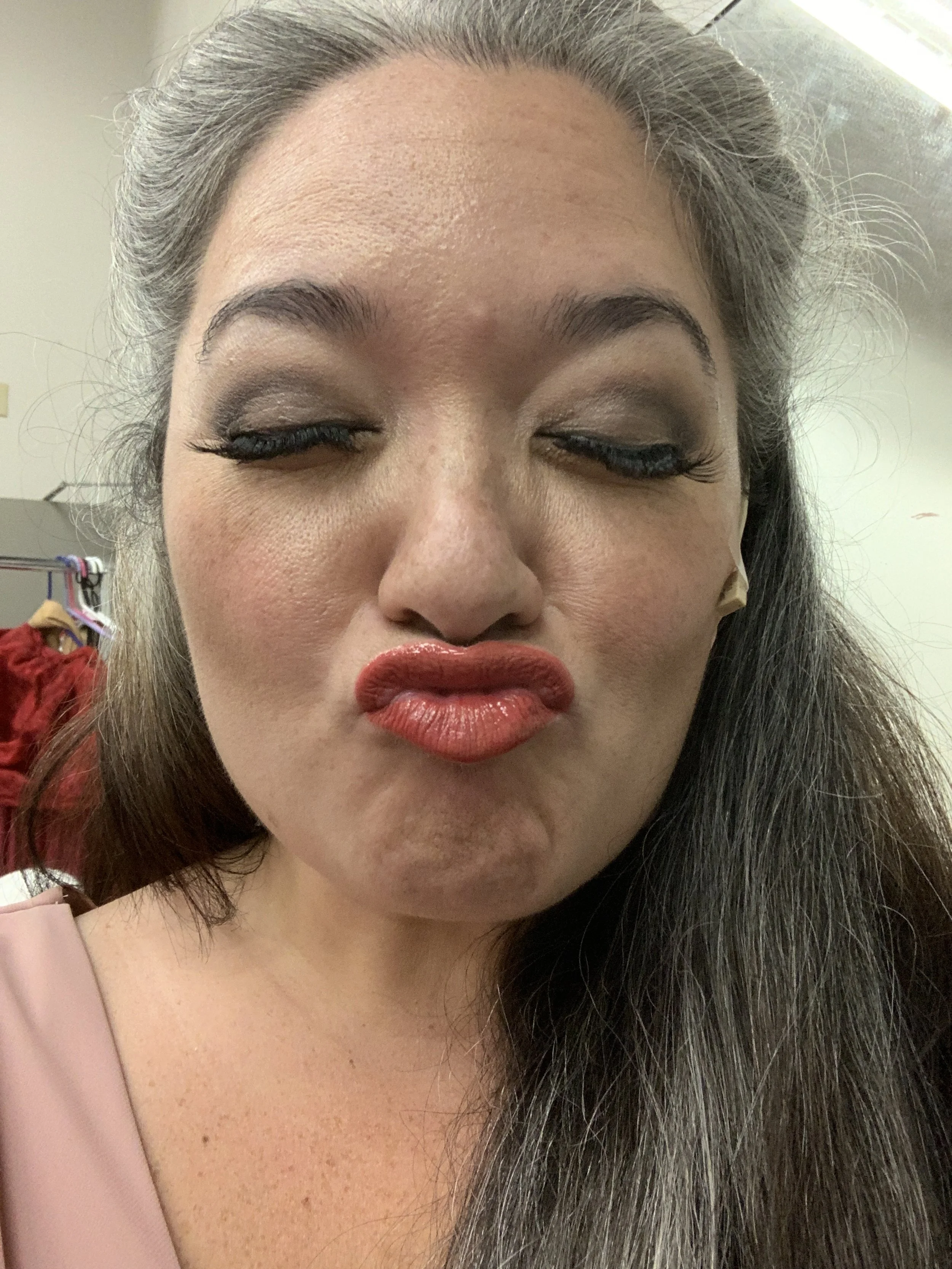 CHILDREN OF EDEN a Broadway musical by I'm A Bright Kid Foundation at Paliku Theatre. Close-up of a woman with makeup, closed eyes, lips puckered in a kiss, and long dark hair with some gray strands, in an indoor setting.