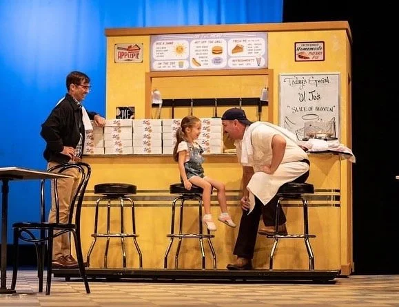 WAITRESS the Broadway musical by Sara Bareilles at Diamond Head Theatre. Child sitting on a bar stool talking to a man dressed as a chef at a small restaurant or café set with a yellow counter, menu boards, and signs in the background.