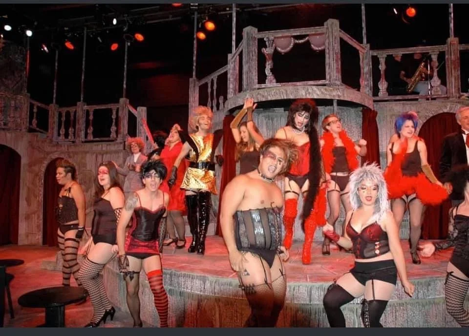 ROCKY HORROR SHOW, Manoa Valley Theatre. A lively stage performance with actors dressed in burlesque or cabaret costumes, featuring corsets, stockings, and feather boas, on a decorated theater stage with wooden balcony railings and colorful lighting.