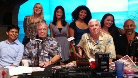 LITTLE SHOP OF HORRORS a comedic musical at Manoa Valley Theatre in Honolulu, Hawaii. Group of eight performers, including men and women, gathered around a table in a room with a blue background, smiling for the photo. Live radio show.