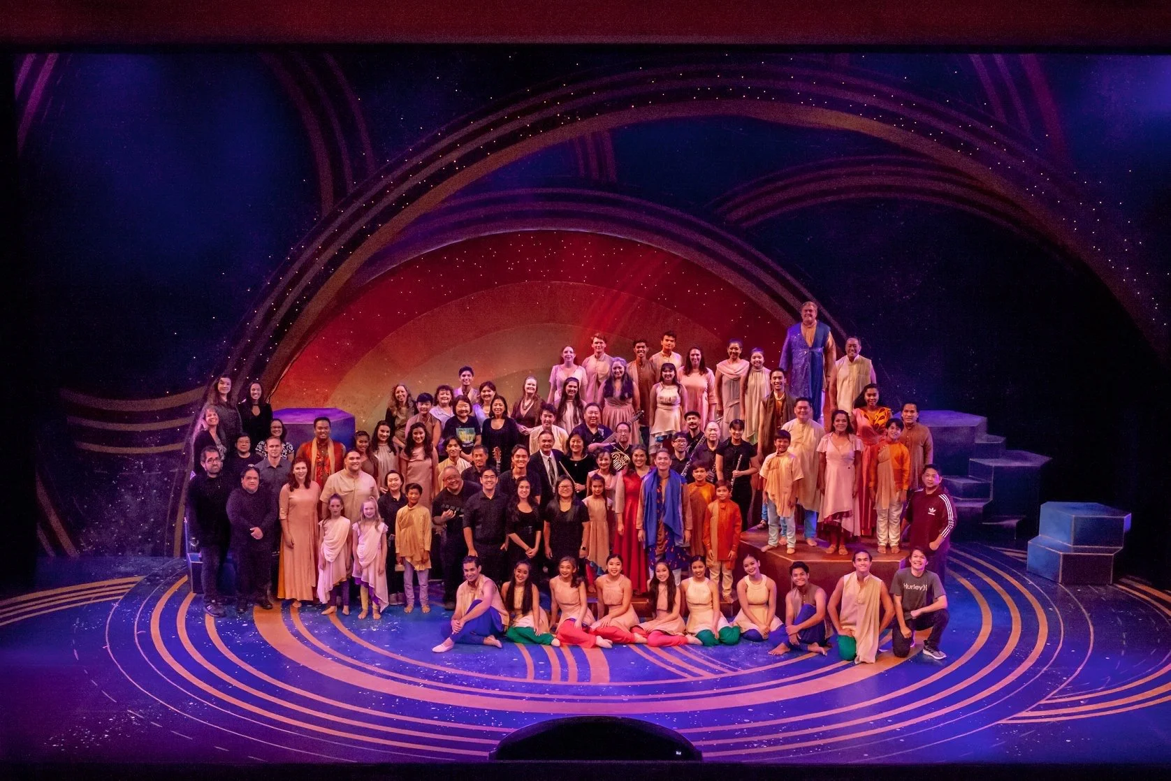 CHILDREN OF EDEN a Broadway musical by I'm A Bright Kid Foundation at Paliku Theatre. A large group of people on a stage, posing for a group photo with a colorful, dazzling backdrop with swirling patterns and stars.