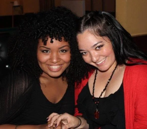 LITTLE SHOP OF HORRORS a comedic musical at Manoa Valley Theatre in Honolulu, Hawaii. Chiko's Tavern. Two women smiling and sitting close together, one with curly black hair and the other with straight dark hair, in an indoor setting.