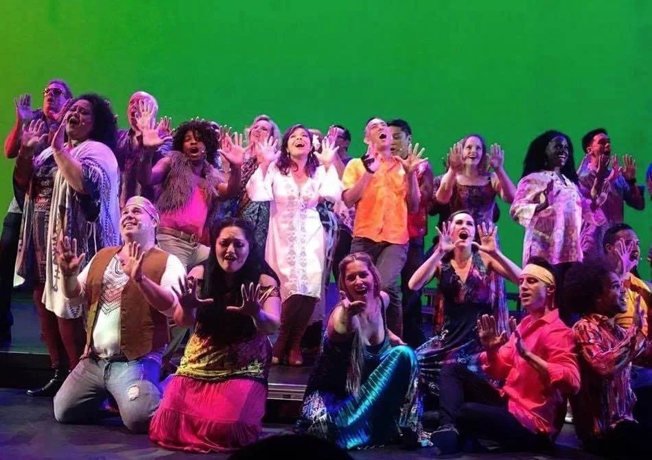 RISE UP: A BENEFIT CONCERT BY MELINA LILLIOS at Paliku Theatre in Kaneohe, Hawaii. Group of diverse people on stage performing with enthusiastic expressions and gestures, under colorful lighting.