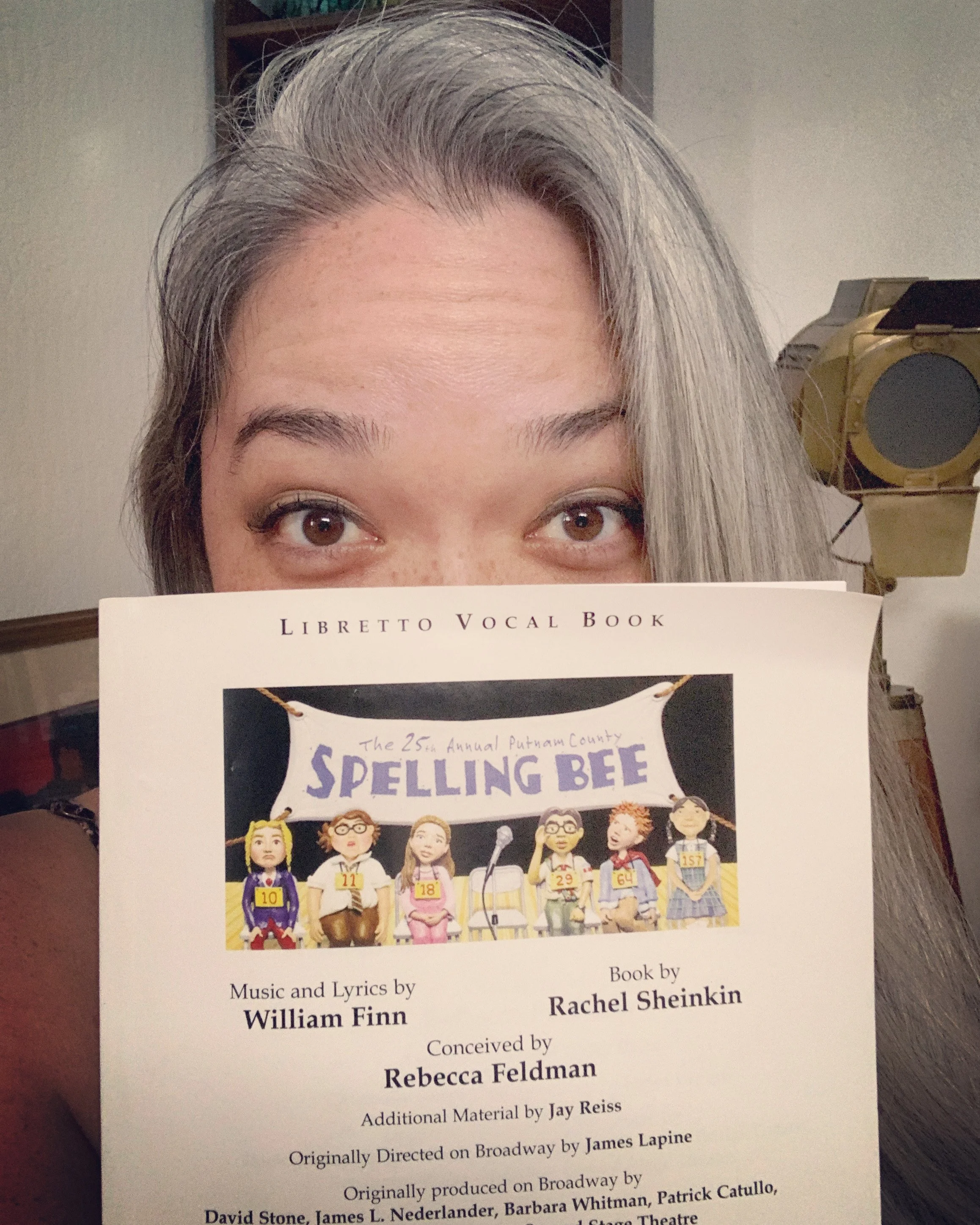 THE 25TH ANNUAL PUTNAM. A person with gray hair holding a script in front of their face, revealing only their eyes and forehead. The script is titled 'Libretto Vocal Book' and features an illustration of animated characters at a spelling bee event.