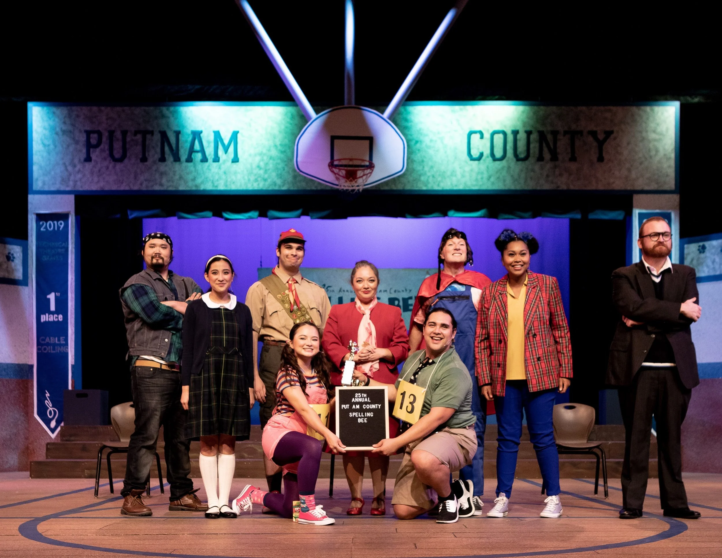 THE 25TH ANNUAL PUTNAM COUNTY SPELLING BEE a comedy. Actors on a stage at a spelling bee event, with a sign reading '25th Annual Putnam County Spelling Bee', in a school gymnasium with a basketball hoop and 'Putnam County' sign overhead.