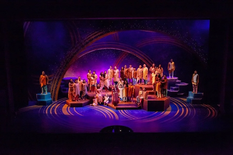 Stage scene with a group of performers in colorful costumes, standing and sitting on tiered platforms, with purple and blue lighting and star-like background.