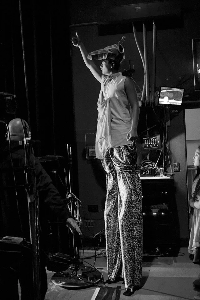 A performer is standing on stilts, wearing a costume with loose, animal print pants and a patterned hat, giving a thumbs-up gesture in a performative setting.