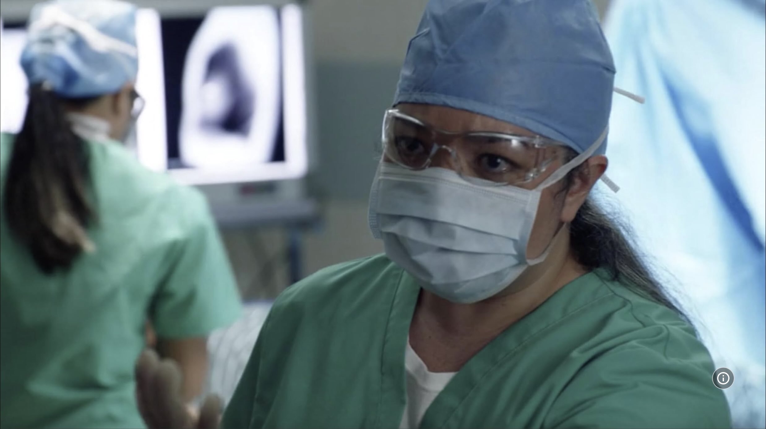 MAGNUM P.I. episode with Cassie Favreau-Chung as a co-star in her recurring role as a nurse in Hawaii. A team of surgeons in green scrubs, wearing masks and glasses, working in an operating room with medical imaging on screens in the background.