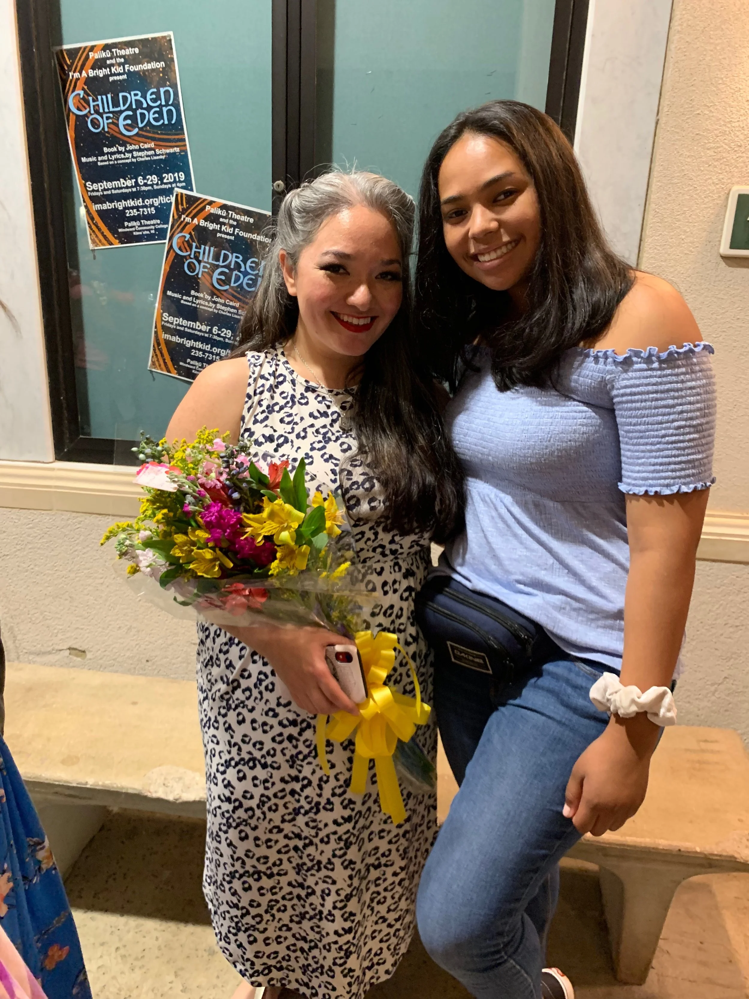 Two women standing close together, smiling. The woman on the left has long dark hair, is wearing a leopard print dress, and is holding a bouquet of colorful flowers. The woman on the right has shoulder-length dark hair, is wearing a light blue off-sh
